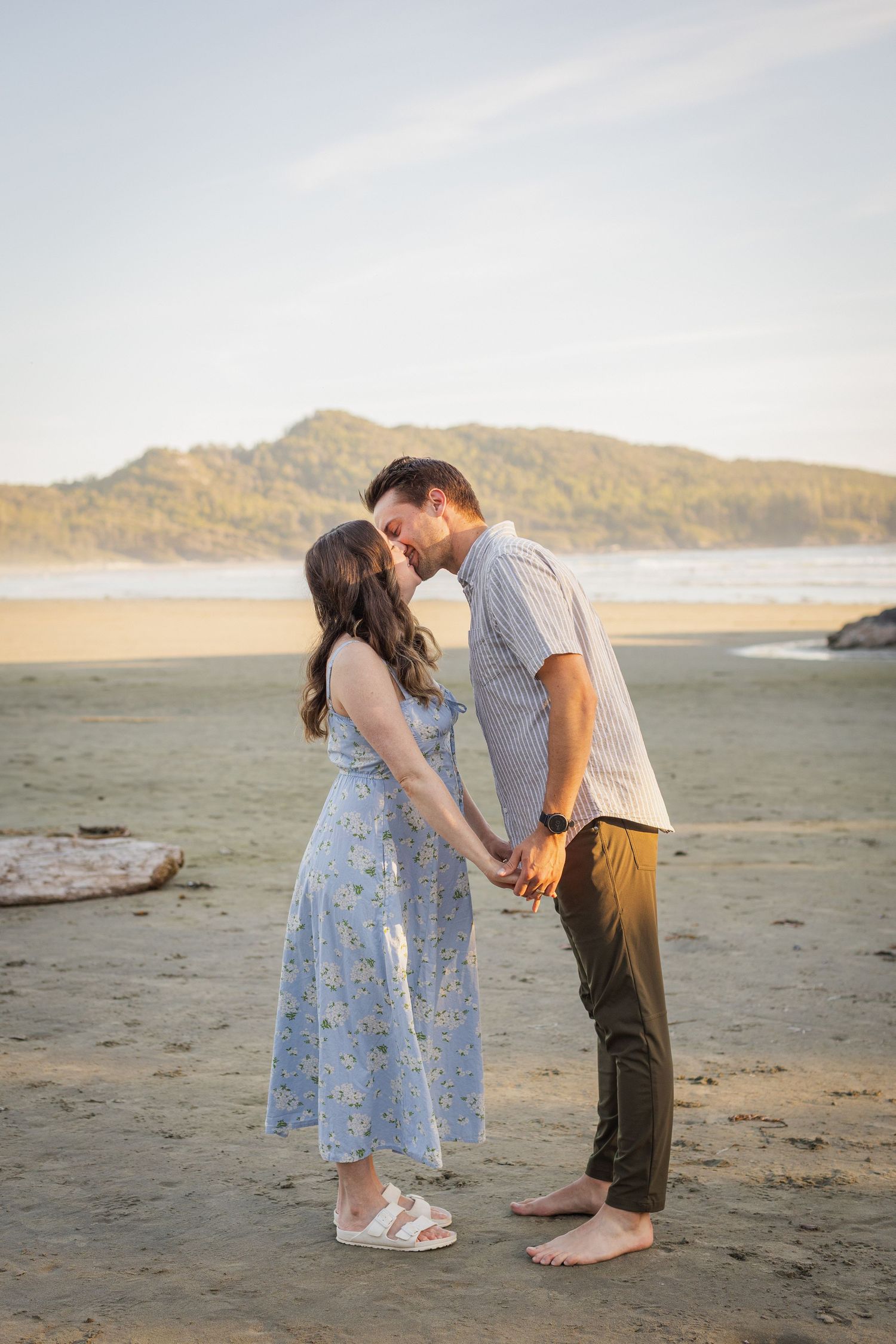 Couple sharing a tender kiss while holding hands on a sandy beach at golden hour with ocean and hills behind.