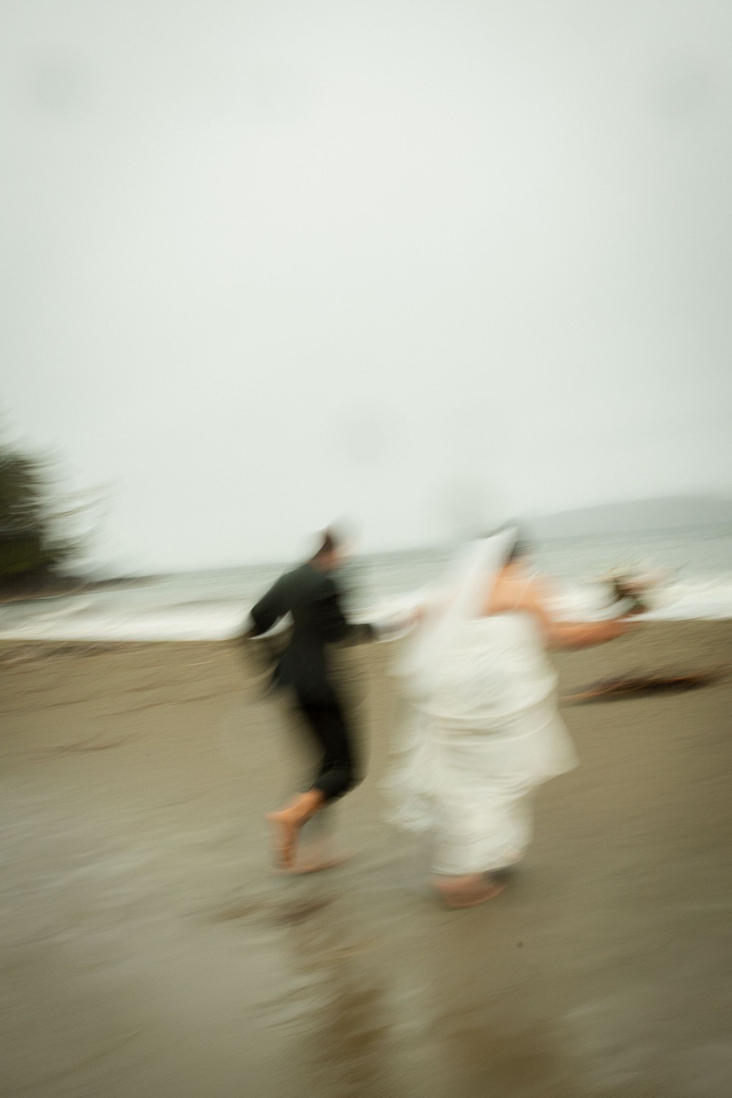 Blurred motion shot of bride and groom running barefoot along a misty beach on their wedding day.