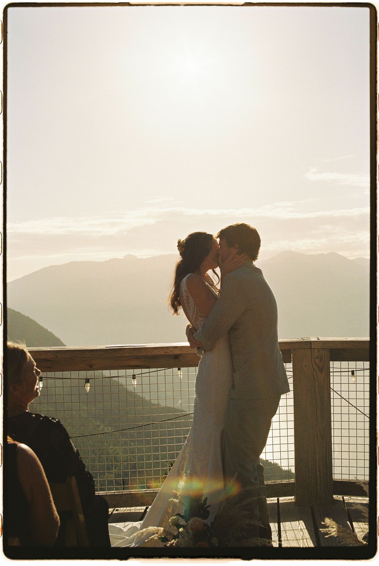 Bride and groom share first kiss silhouetted against golden sunset sky with mountain range backdrop.