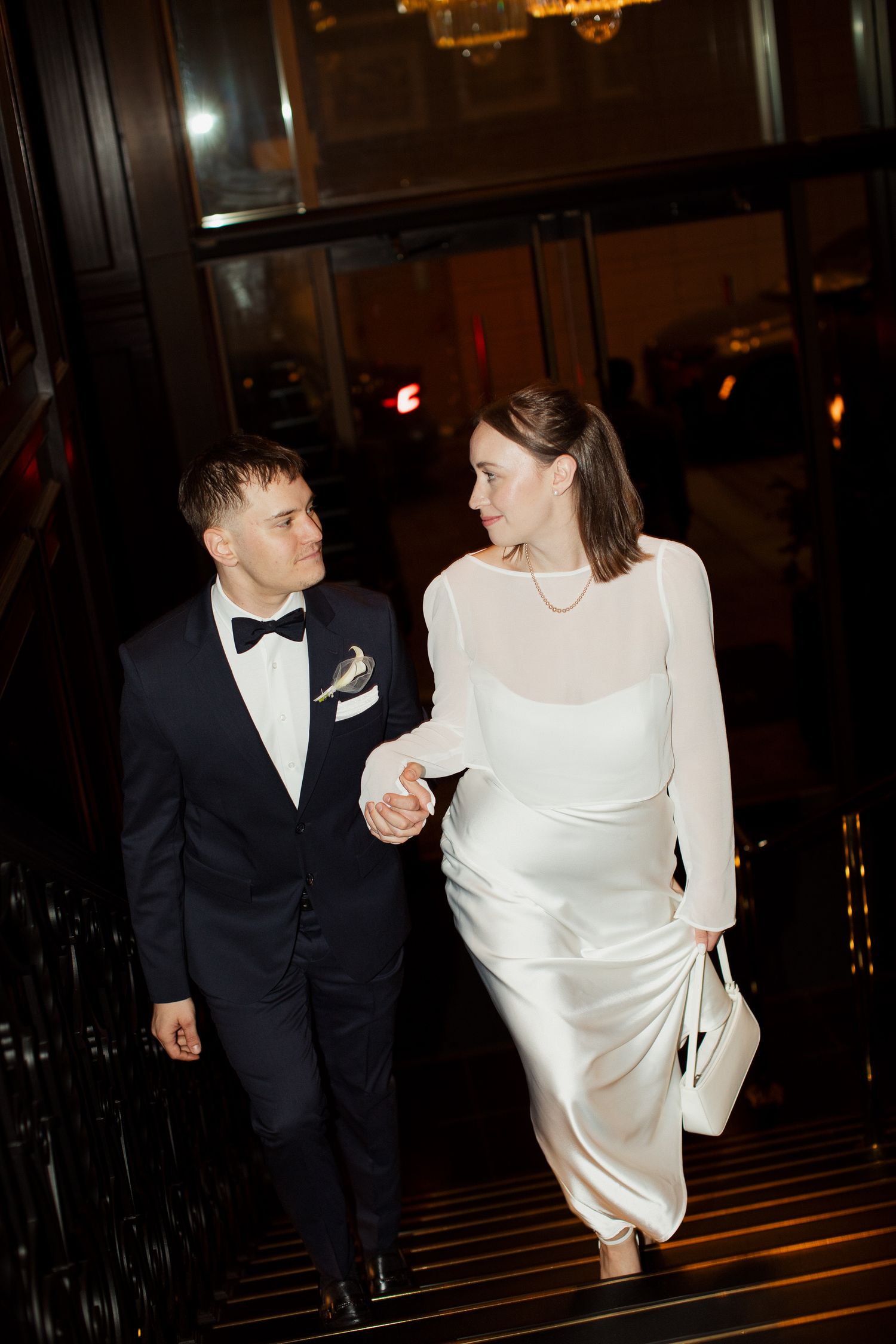 Couple in formal wedding attire holding hands while descending grand staircase.