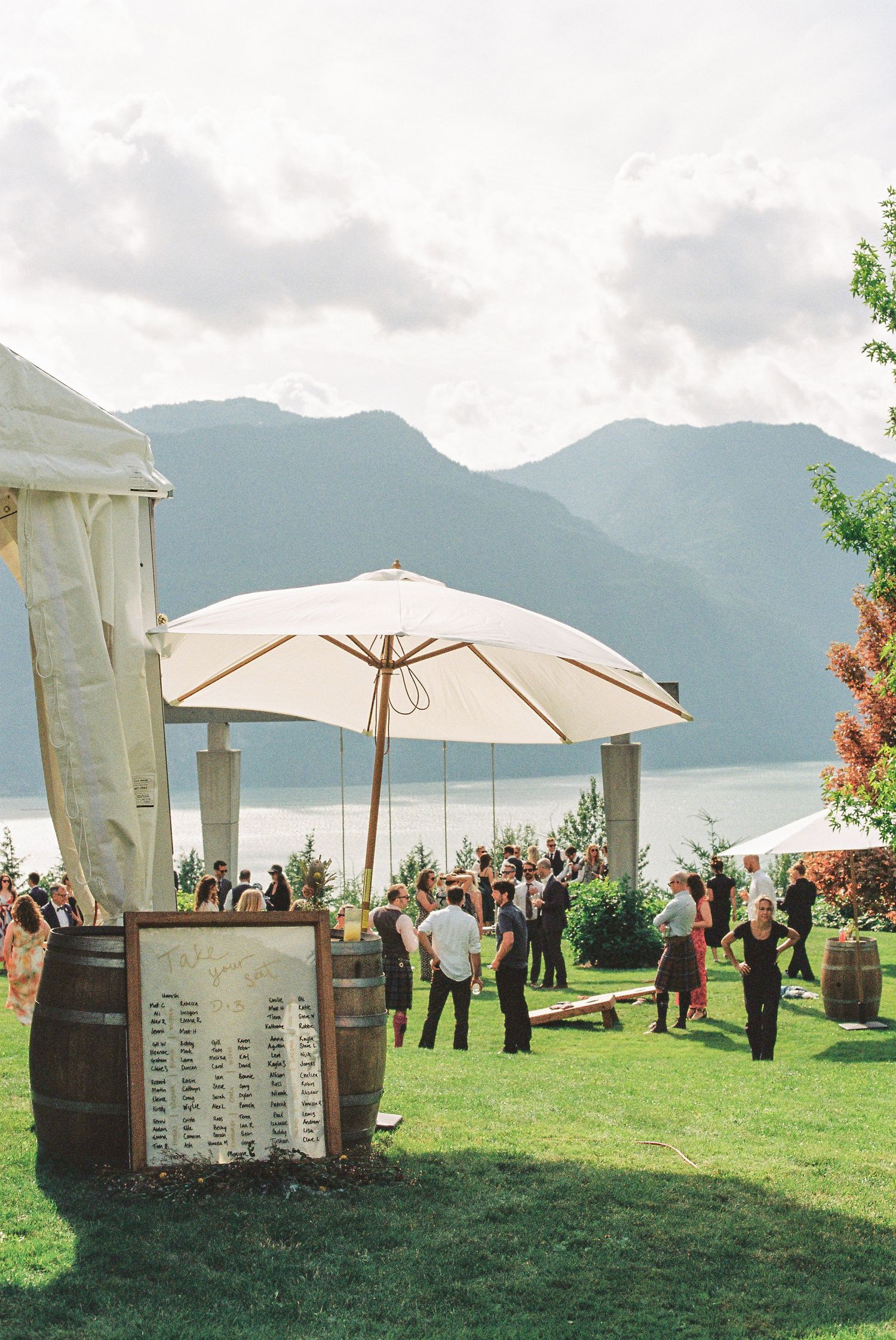 Outdoor wedding cocktail hour on lawn with mountain lake views, barrel seating chart, and white umbrellas.