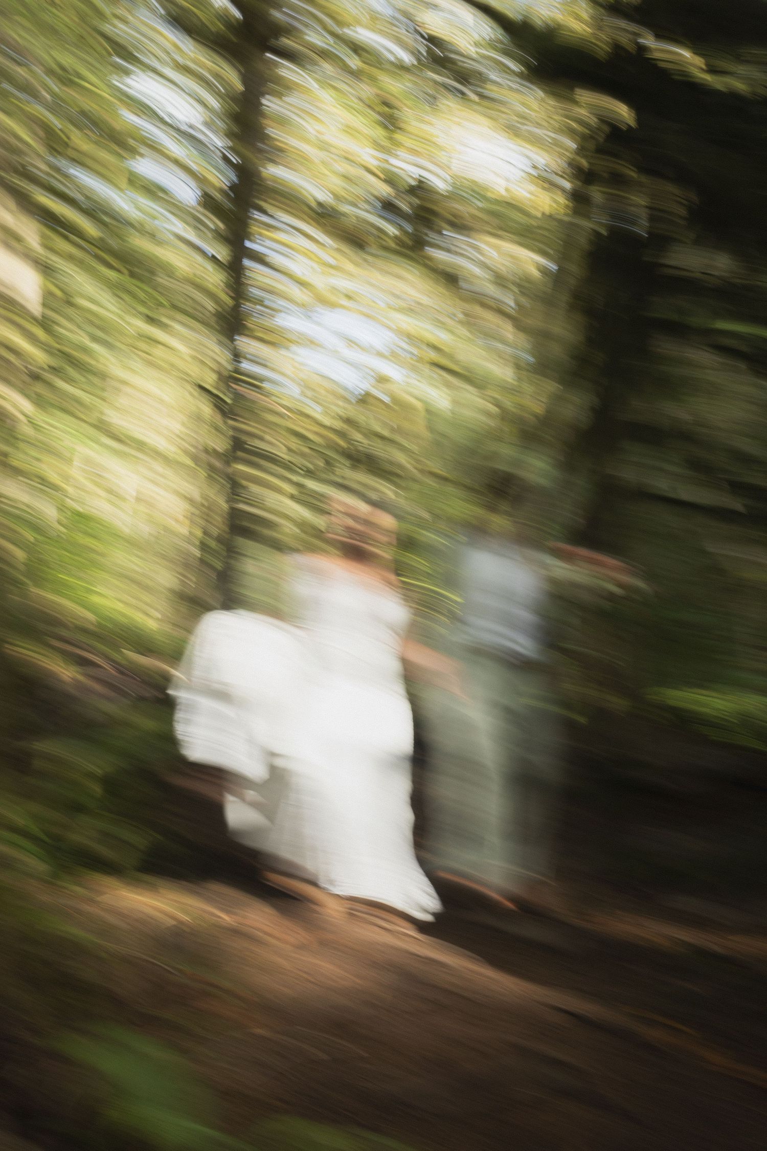 Blurred motion shot of bride in white dress walking through lush green forest.