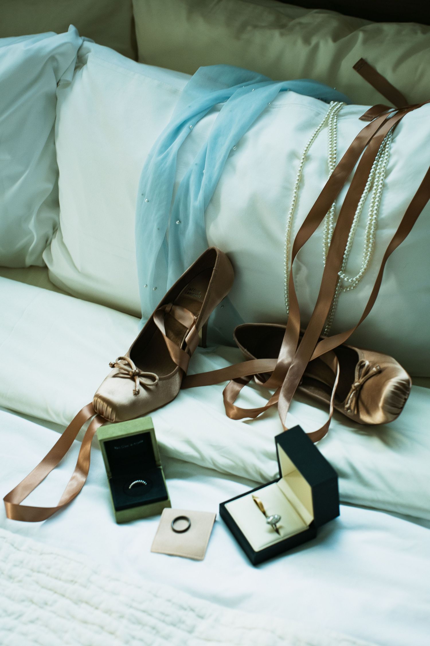 Gold satin bridal shoes with ribbon ties beside wedding rings in boxes and pearl necklace on bed.