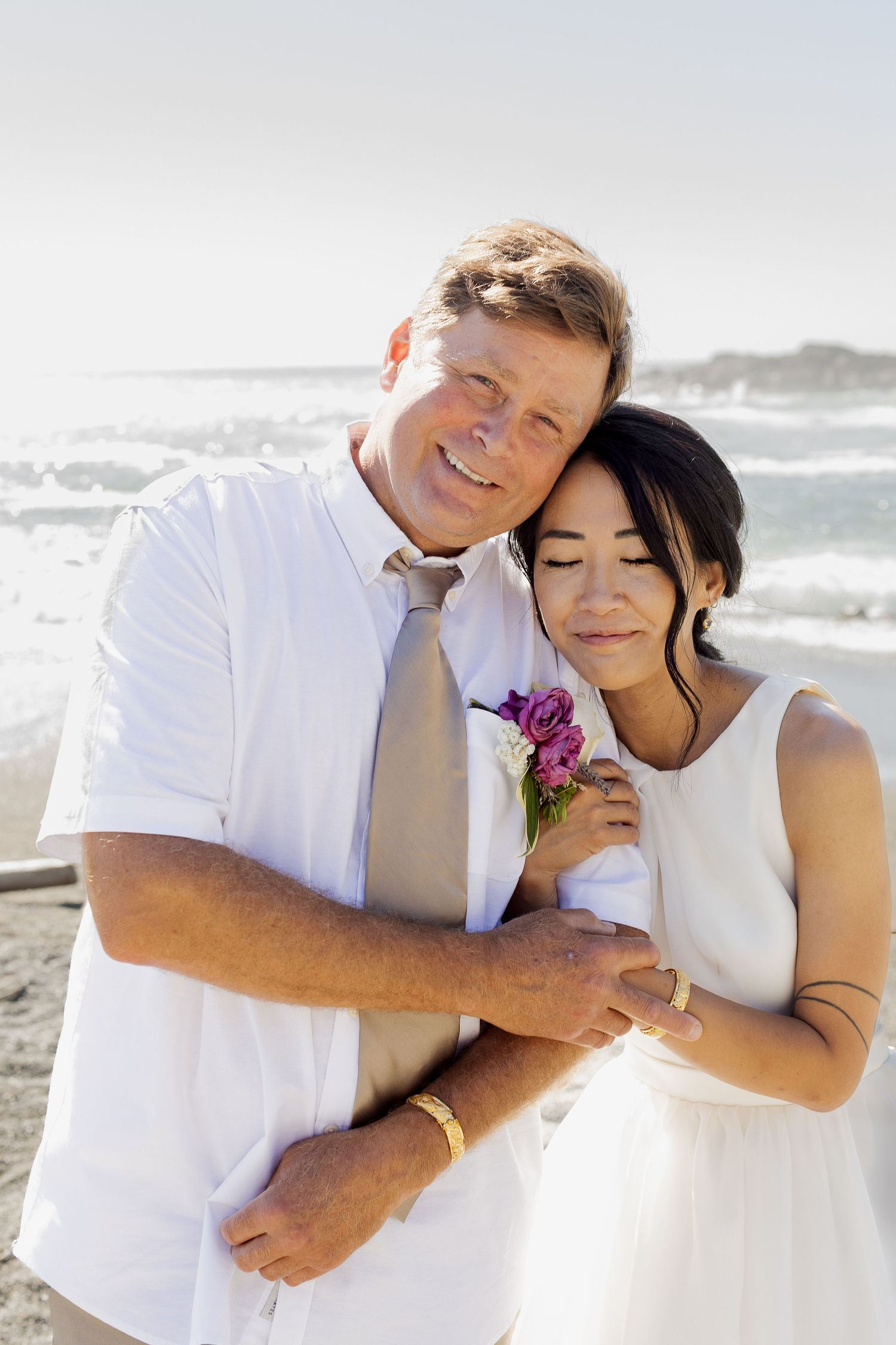 Happy couple embracing on sunny beach, bride holding purple flowers, groom in white shirt and tie.