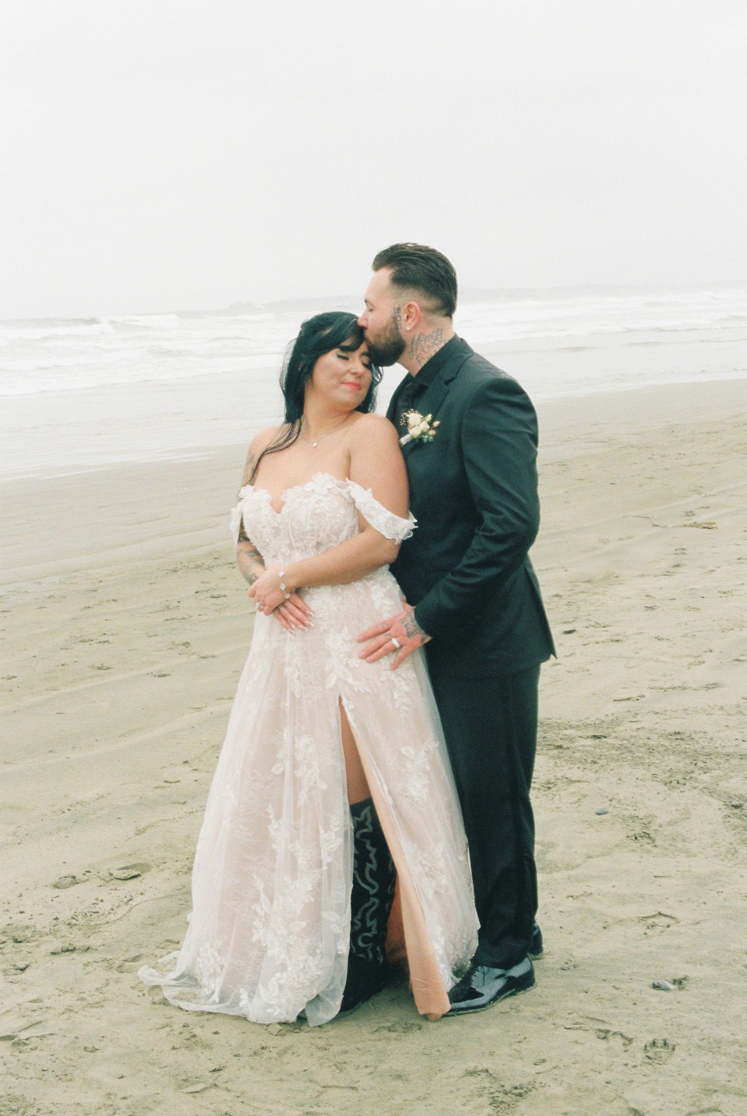 Bride in lace gown and groom in dark suit embracing on foggy beach.