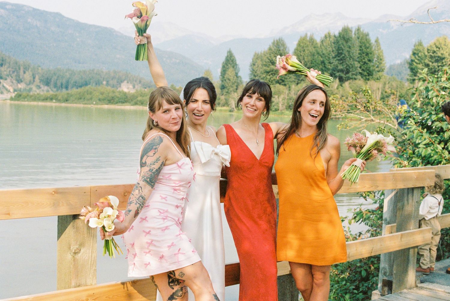 Bride and three bridesmaids in colorful dresses celebrating on wooden deck with mountain lake view.