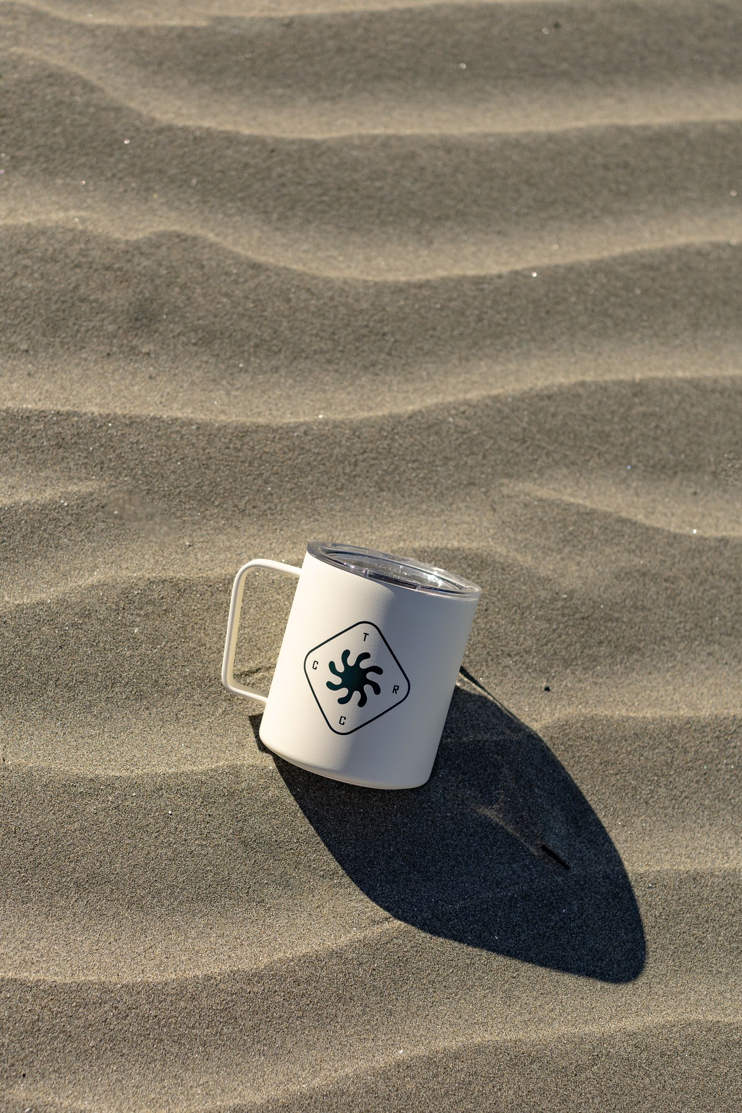 White insulated camp mug with sun logo casting shadow on rippled sandy beach surface.