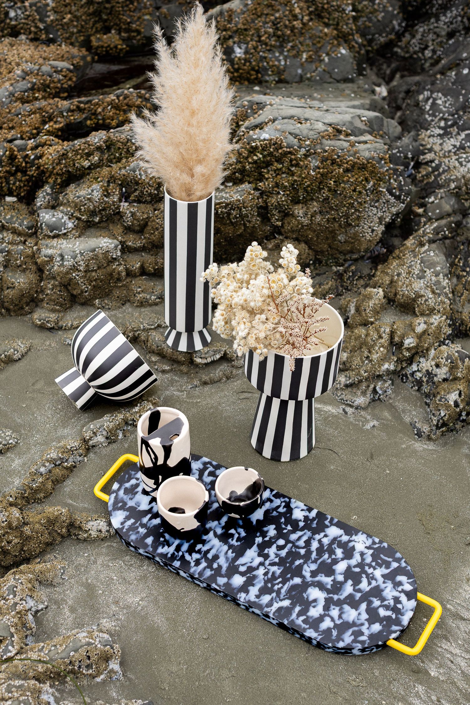 Bold black and white striped vases with pampas grass and spotted ceramics on tray among rocks.