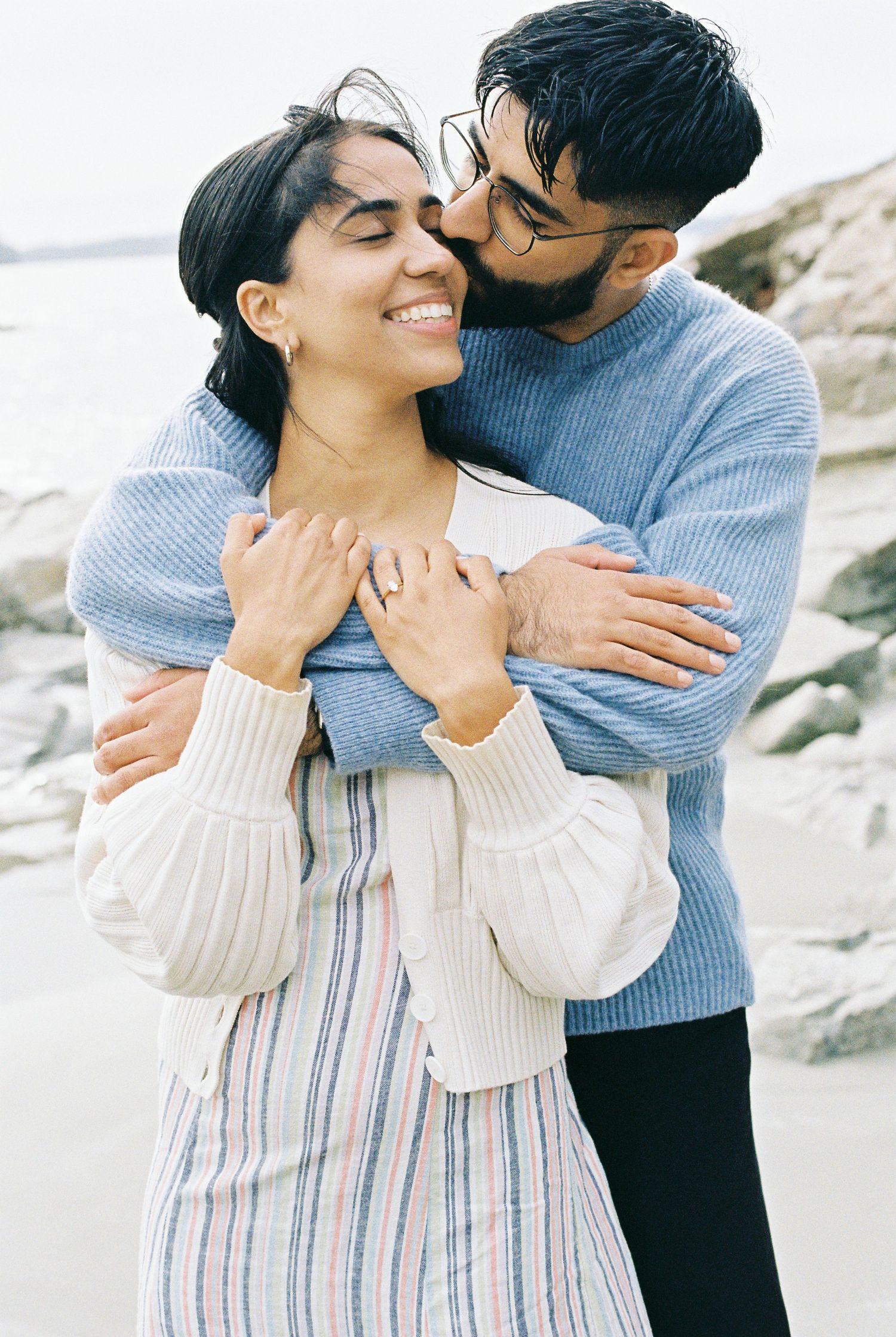 Man hugging woman from behind on rocky beach as she smiles with eyes closed.