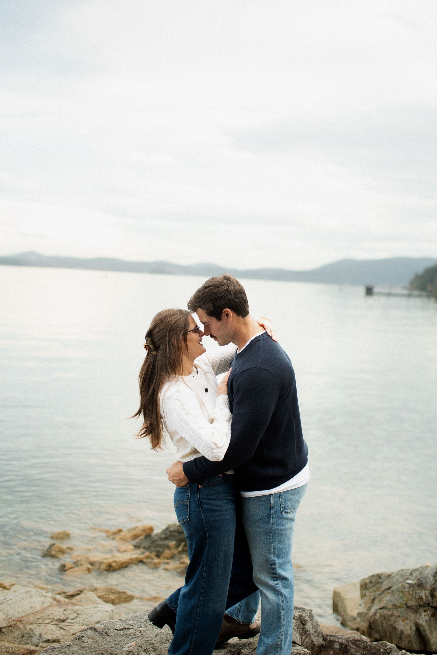 Romantic couple embracing face-to-face on a rocky shoreline with a serene lake backdrop.