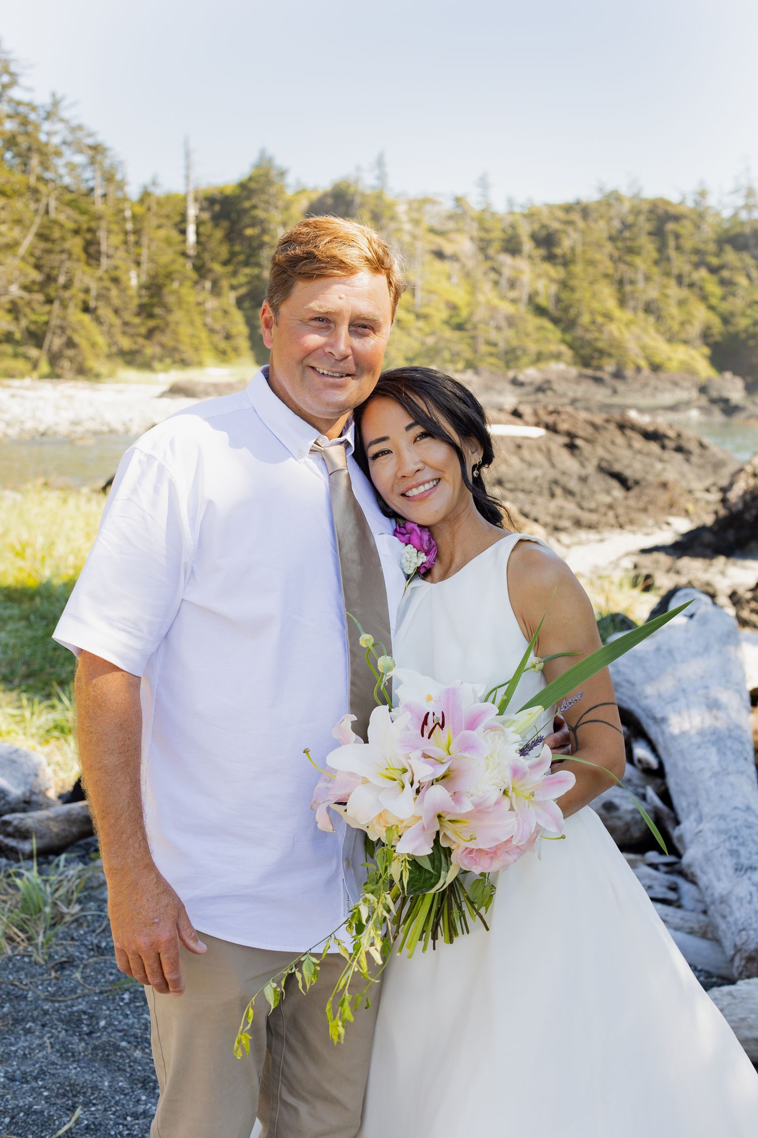 Smiling bride and groom pose together outdoors holding pink flower bouquet near rocky coastline.