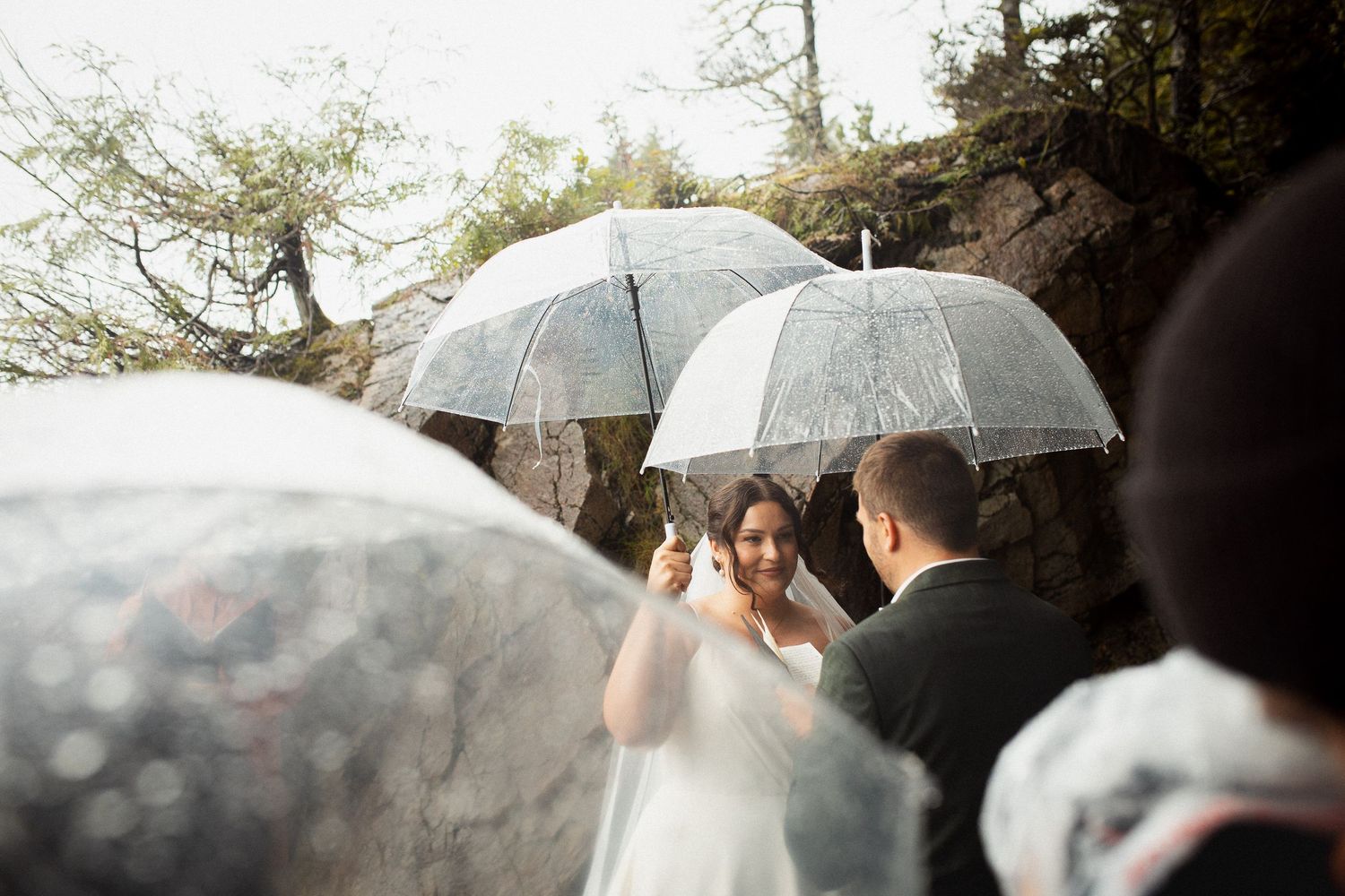 Smiling bride holds bouquet during rainy beach wedding ceremony surrounded by clear umbrellas.