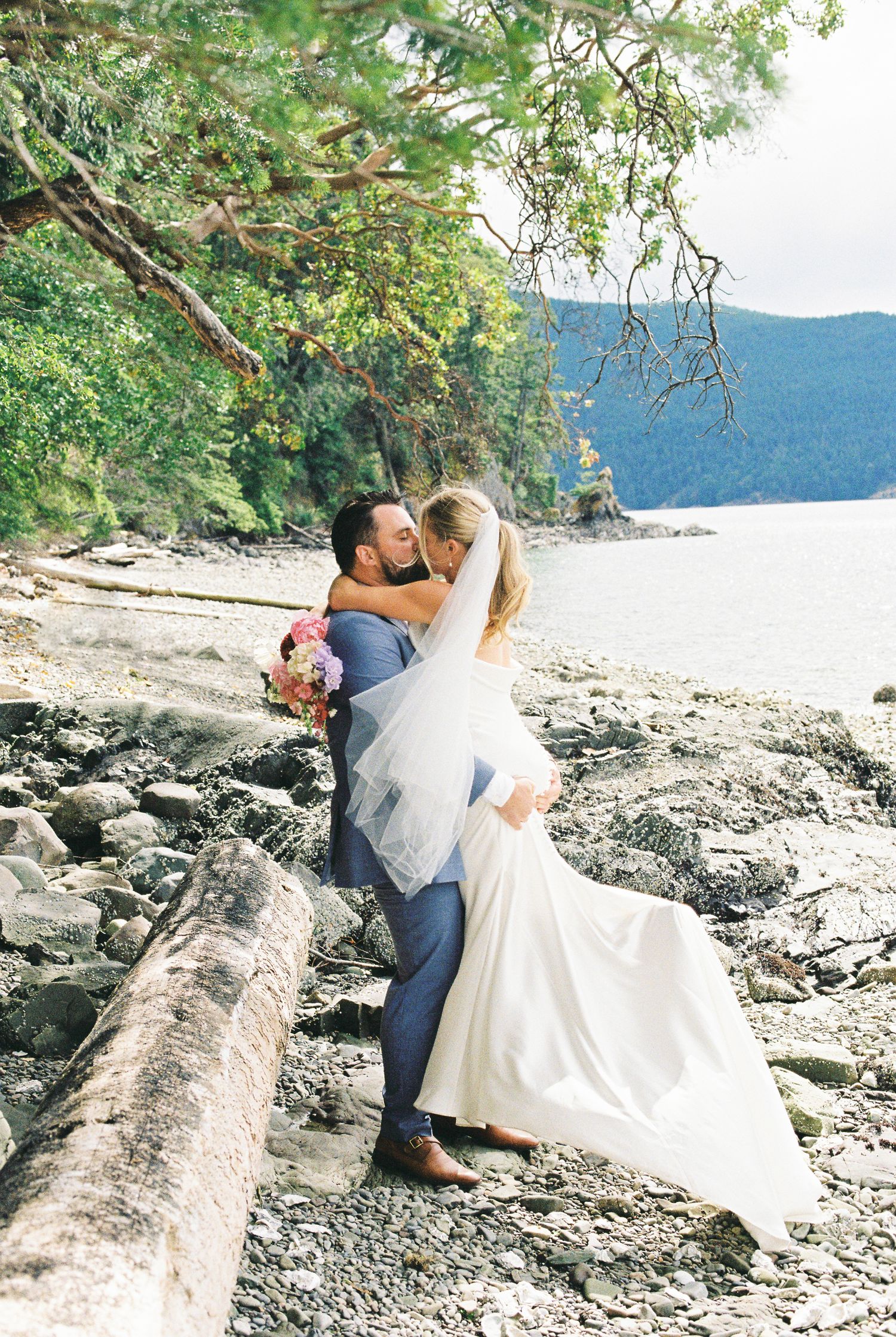 Groom lifting bride in white gown and veil on a rocky beach surrounded by lush green trees.
