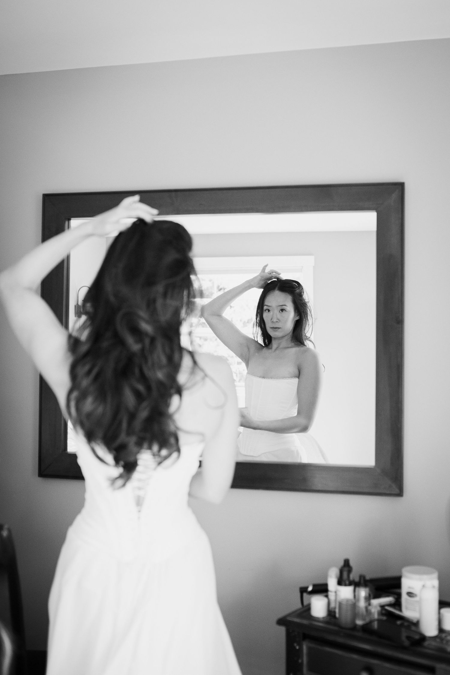 Black and white photo of bride in strapless gown adjusting hair while looking in mirror before wedding.