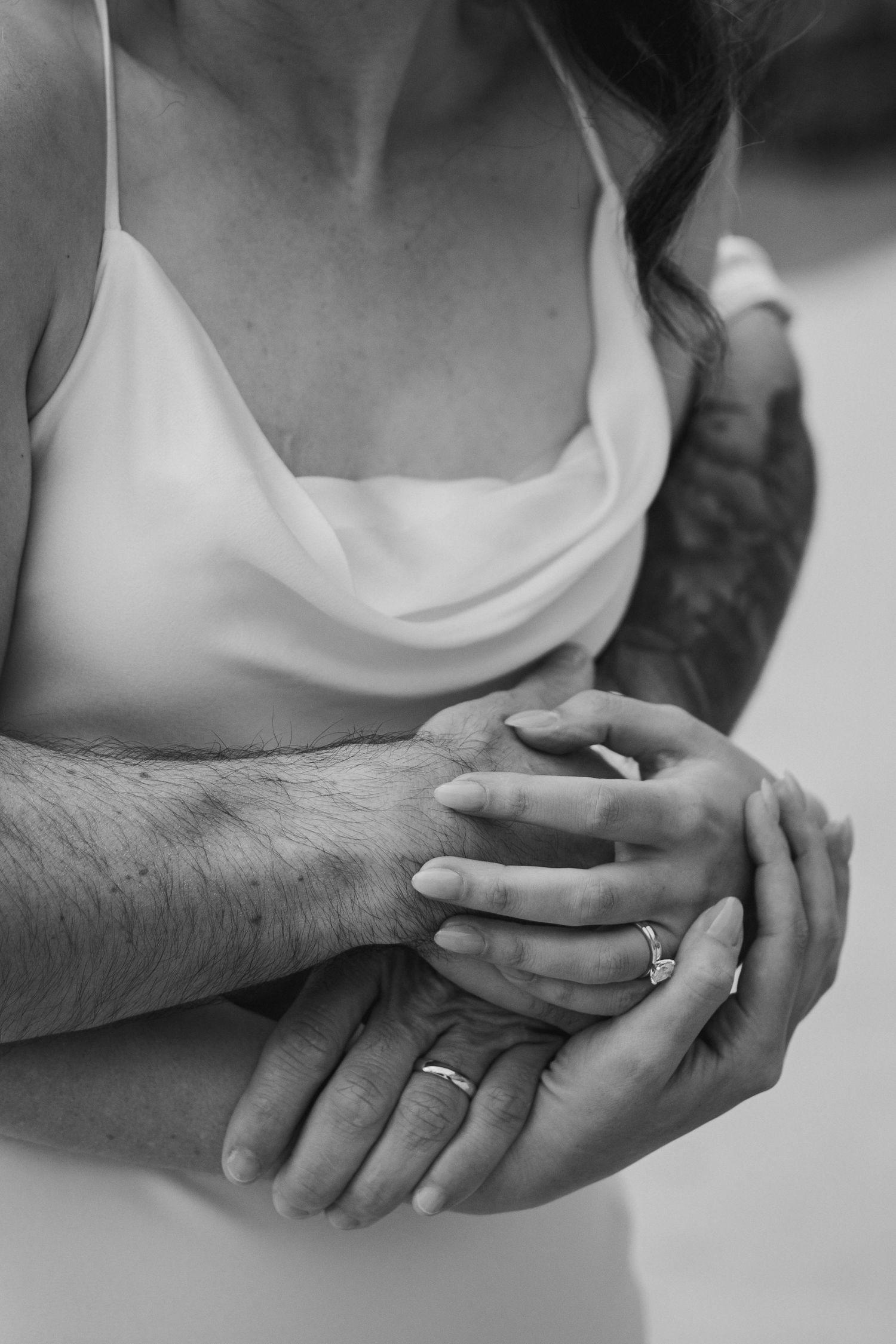 Black and white close-up of couple holding hands, showing engagement ring and wedding band with tattoos.