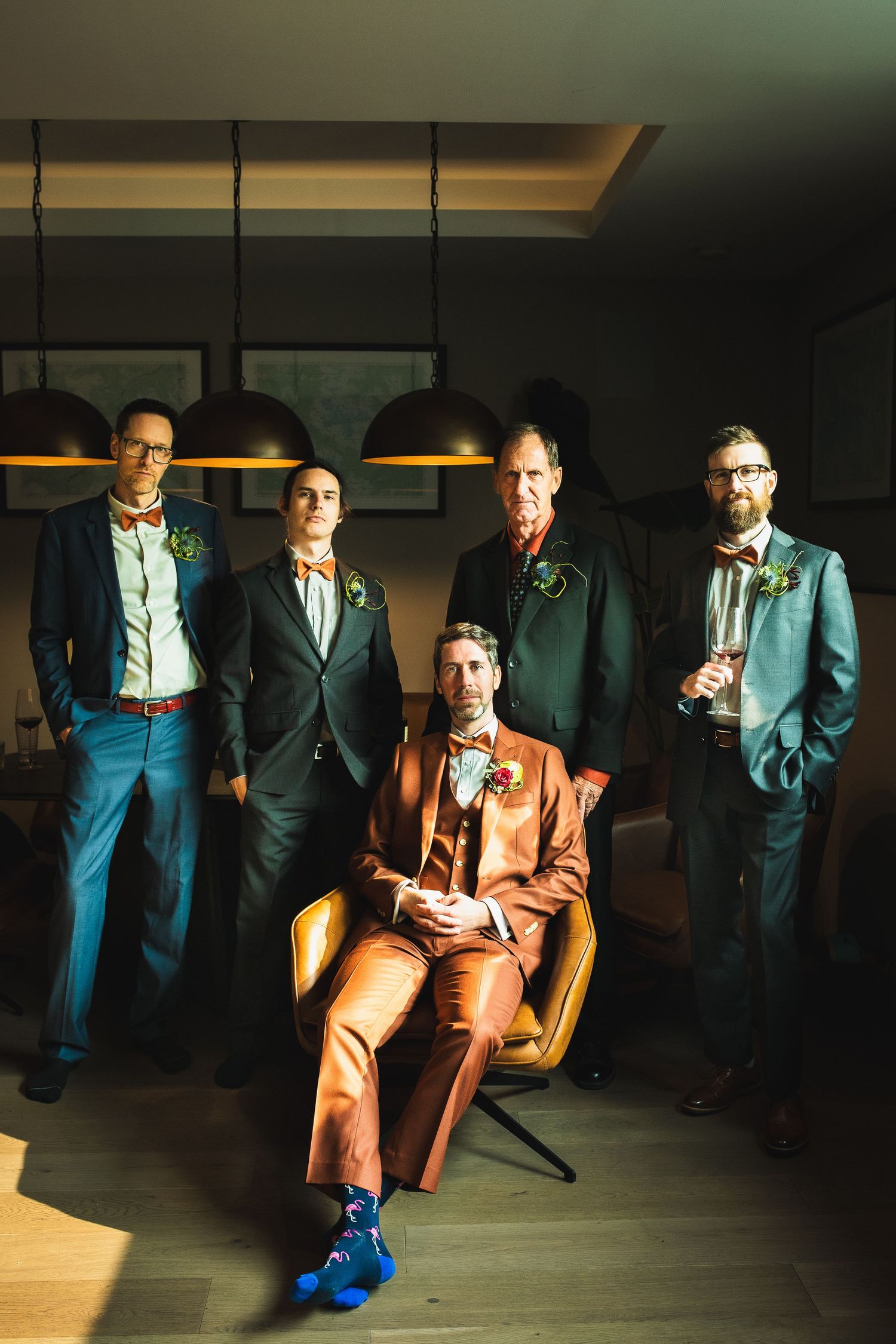 Groom in rust orange suit seated among four groomsmen in dark suits with bow ties indoors.