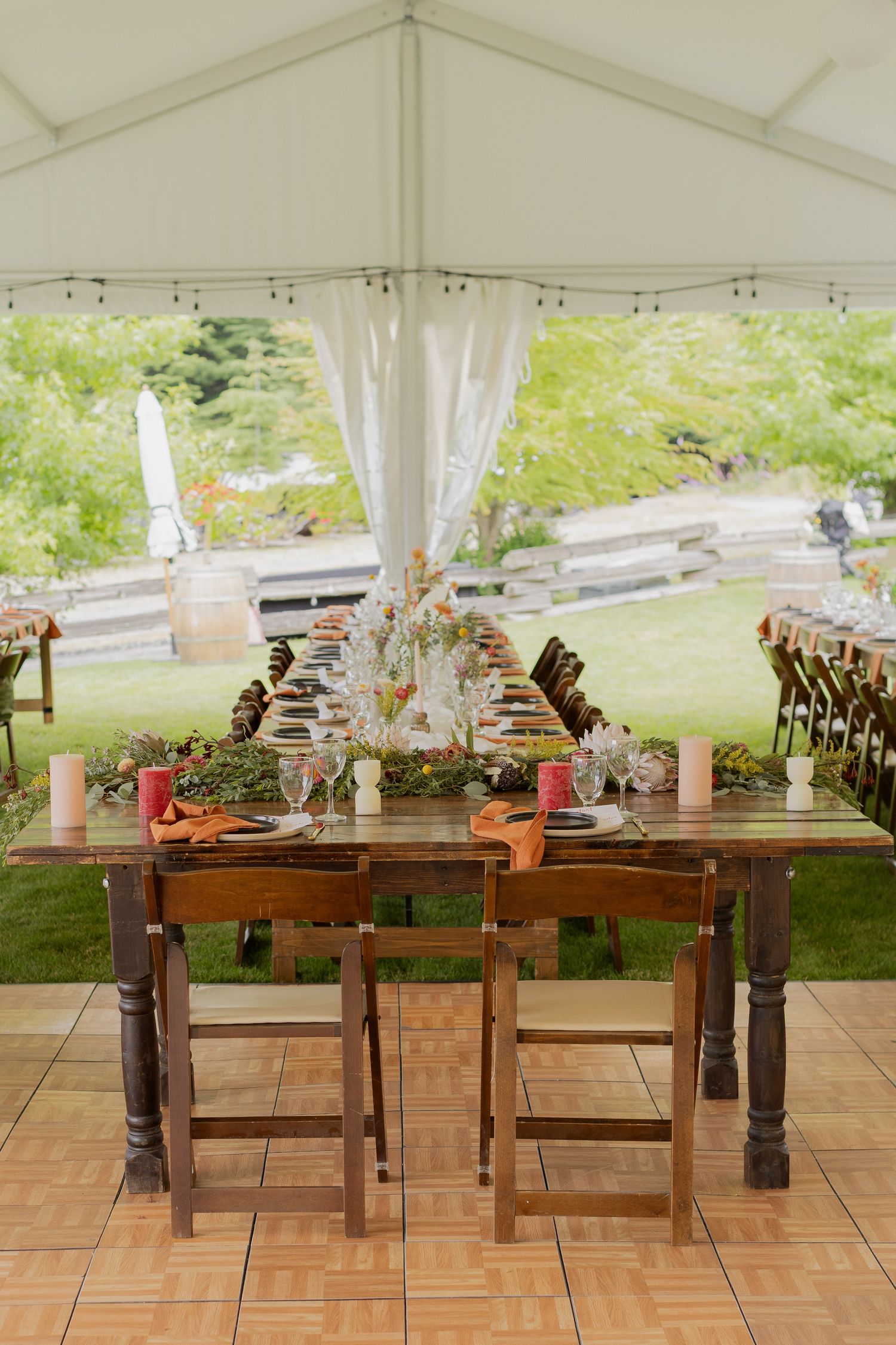 Rustic wooden farm table set with floral centerpieces under white wedding tent.