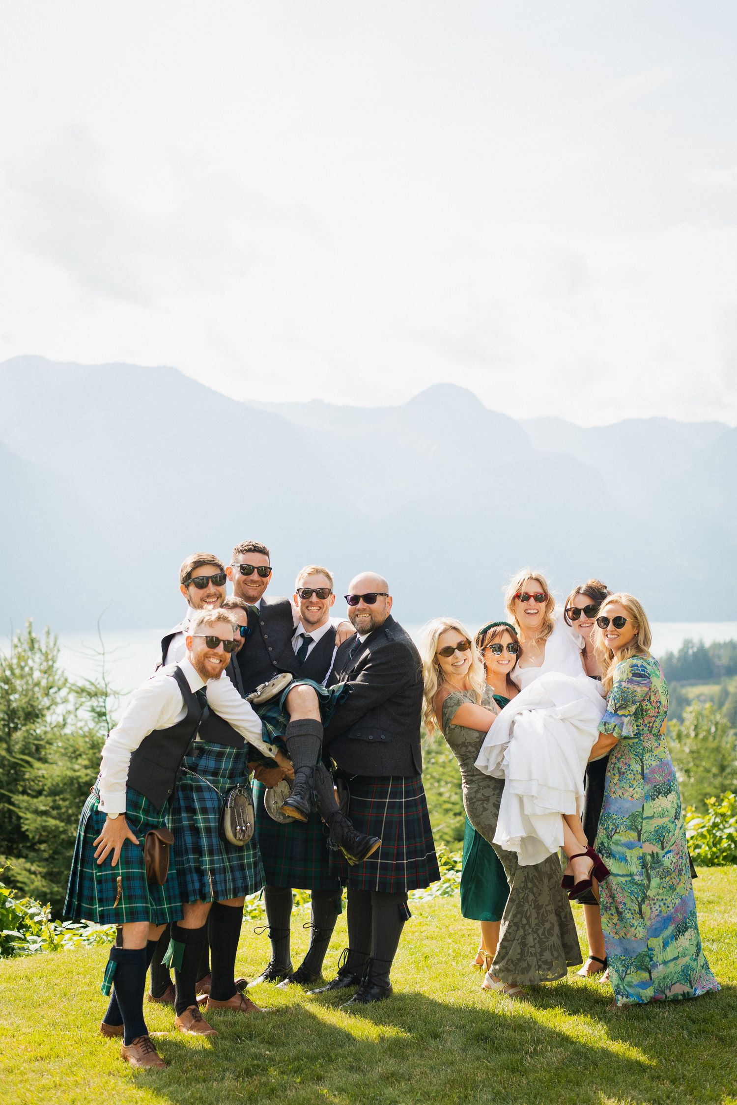 Wedding party in kilts and formal wear posing with bride outdoors against mountain backdrop.