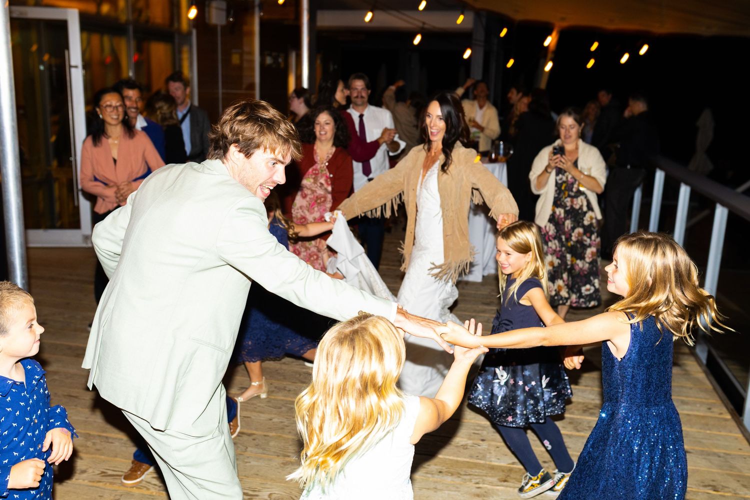 Groom dancing joyfully with children and guests on wooden dance floor at wedding reception.