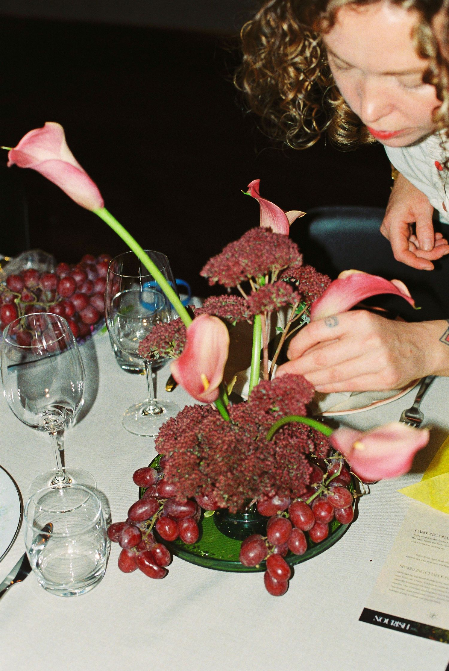 Curly-haired woman arranging pink calla lily and sedum centerpiece with red grapes on white tablecloth.