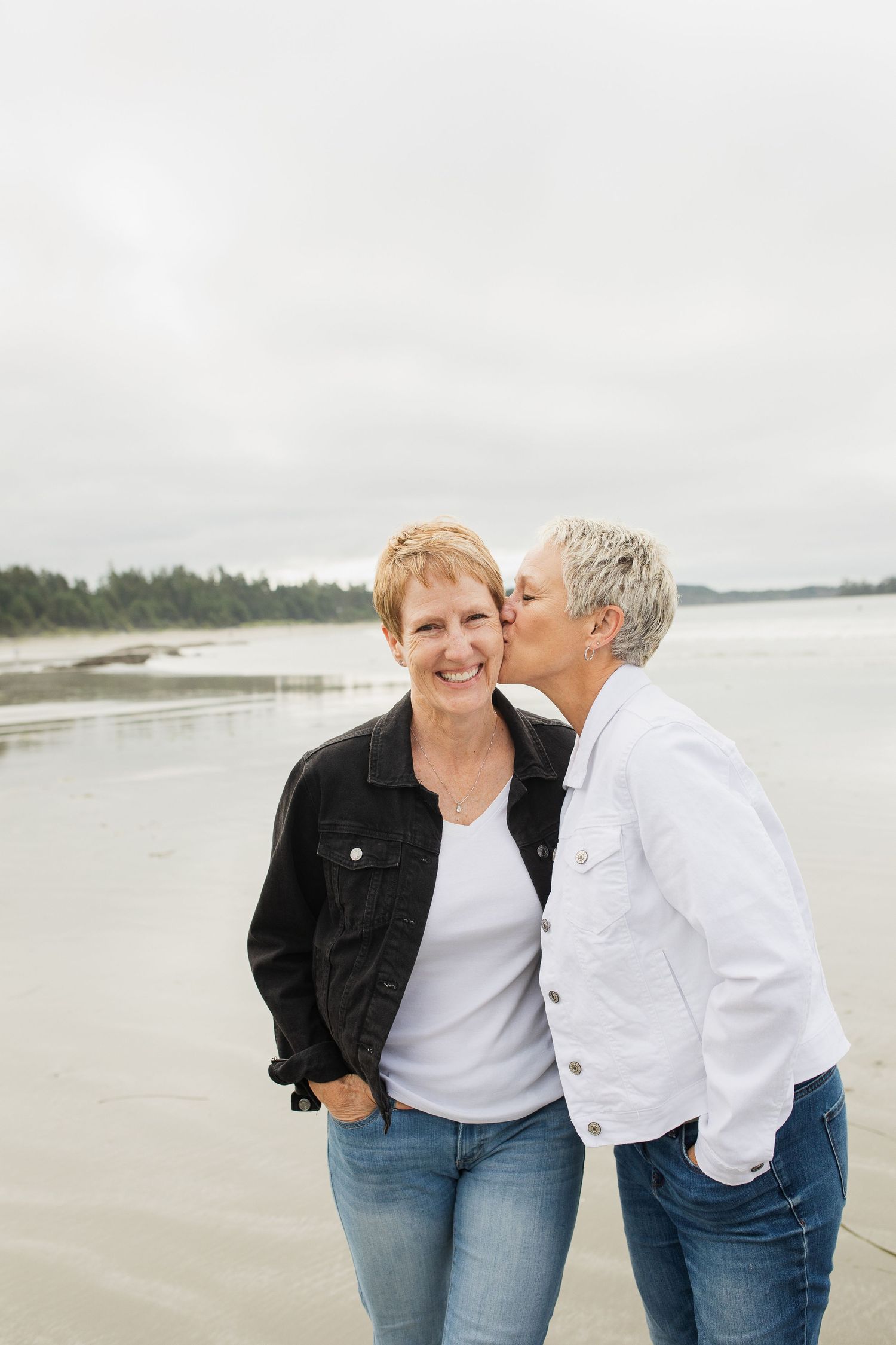 Two mature women smiling on a beach, one kissing the other on the cheek affectionately.