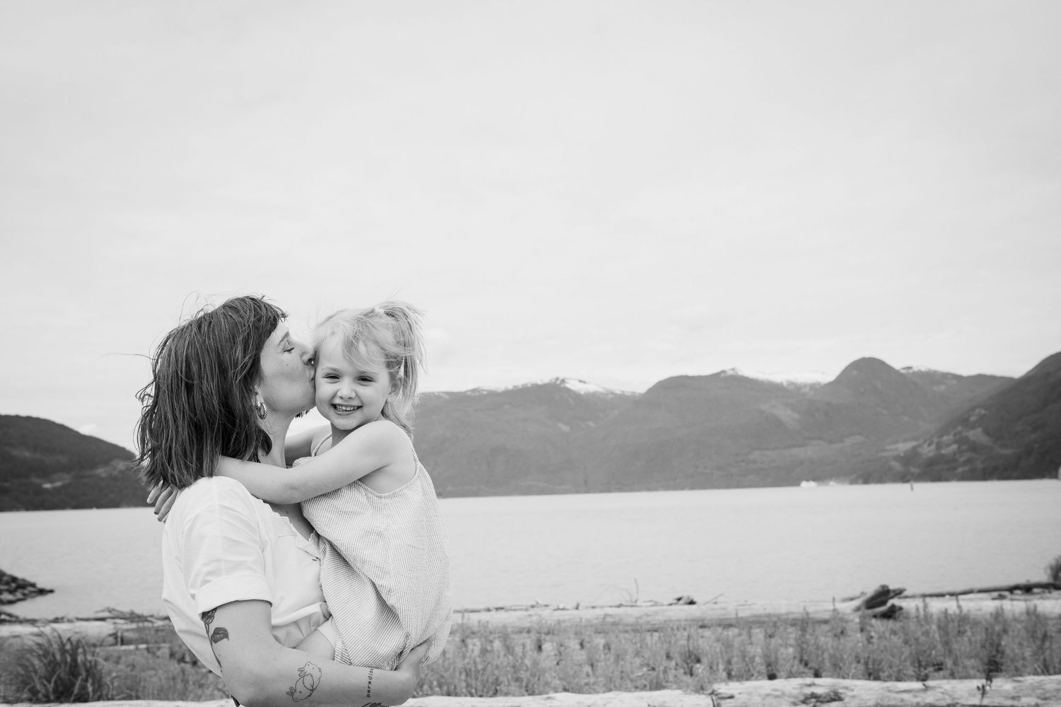 Black and white photo of mother kissing young daughter held in arms near mountain lake.