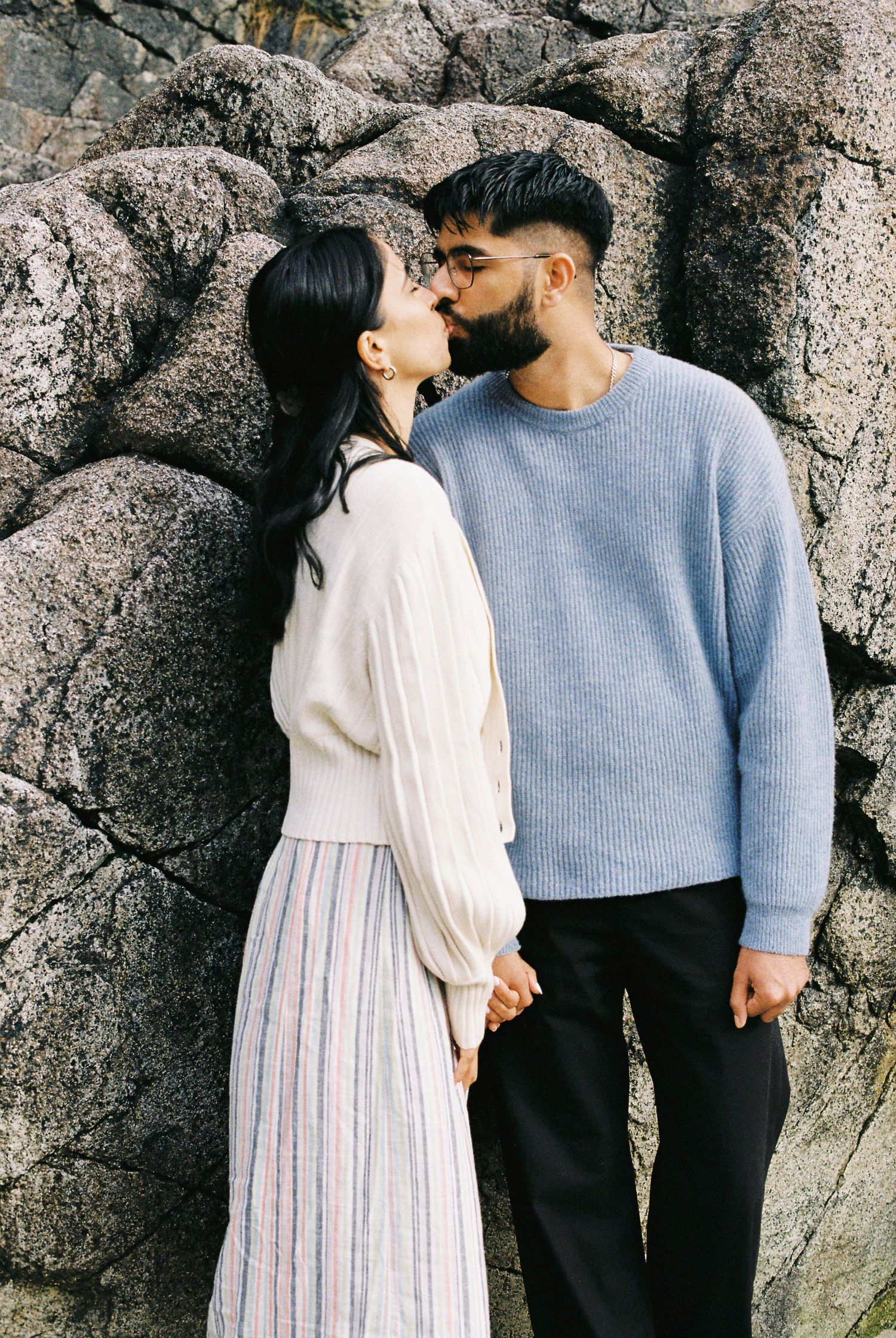 Couple sharing a gentle kiss while holding hands against a textured rock wall.