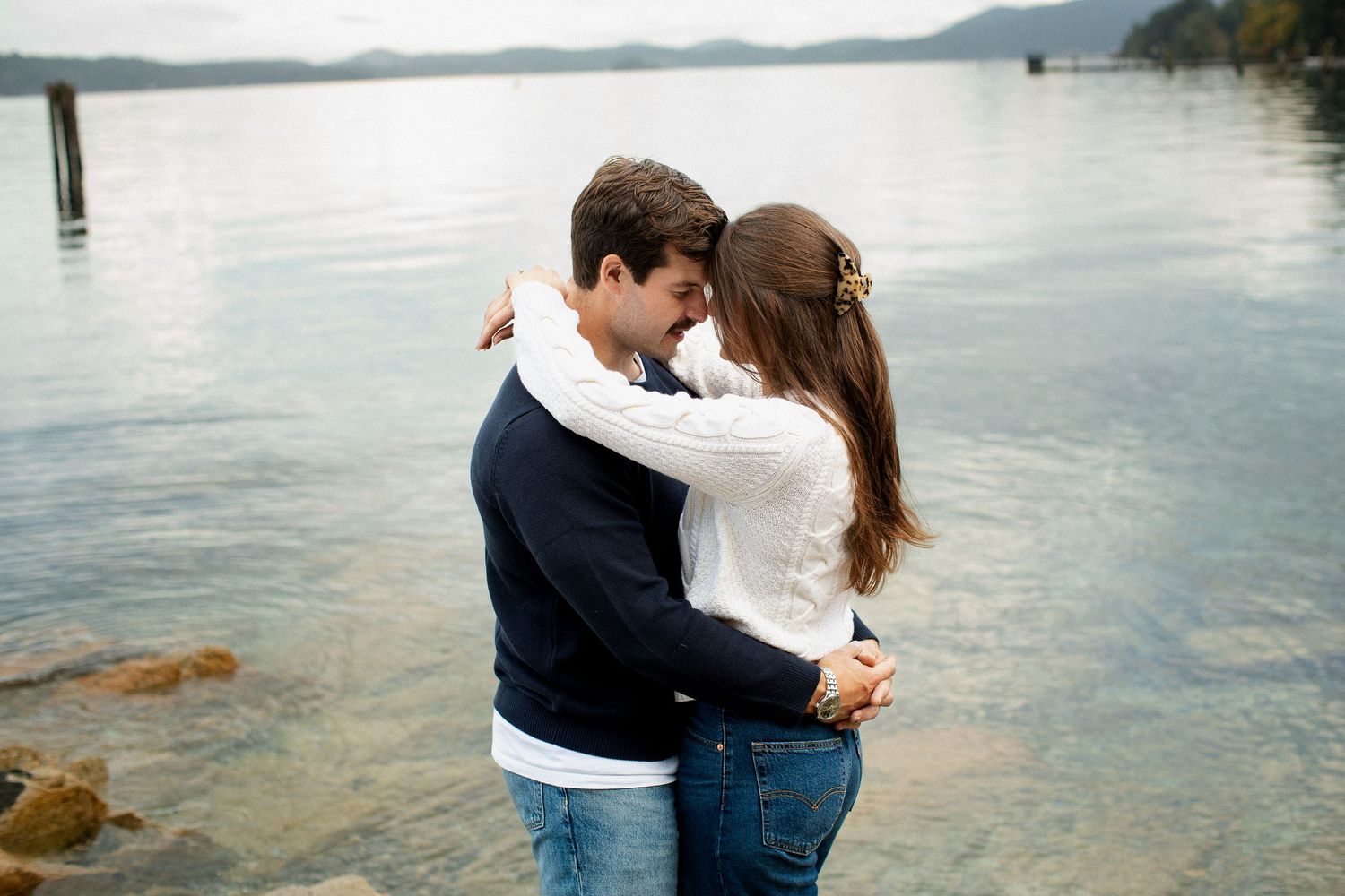 Couple embracing and smiling at each other by a calm lake with mountains in the background.