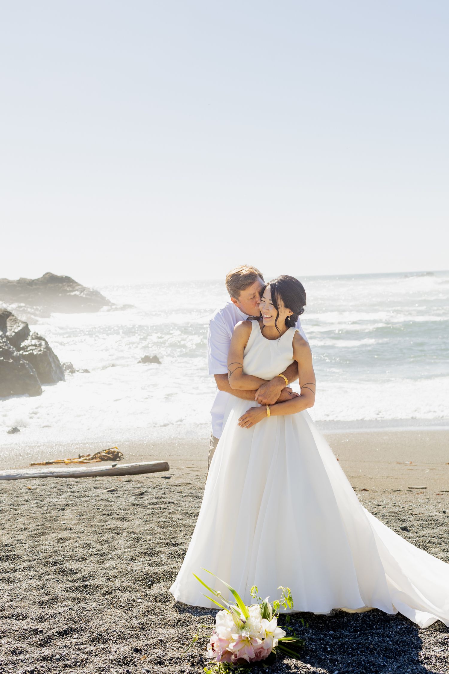 Bride and groom share romantic embrace on sandy beach with ocean waves in background.