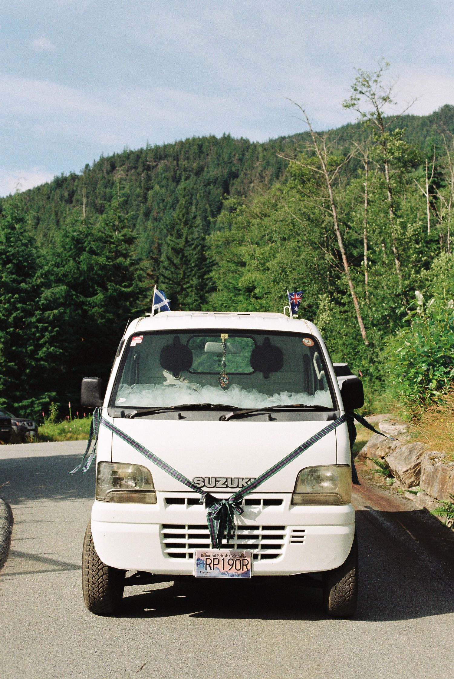 White Suzuki mini truck decorated with ribbon bow and Scottish flag for wedding transportation in mountains.