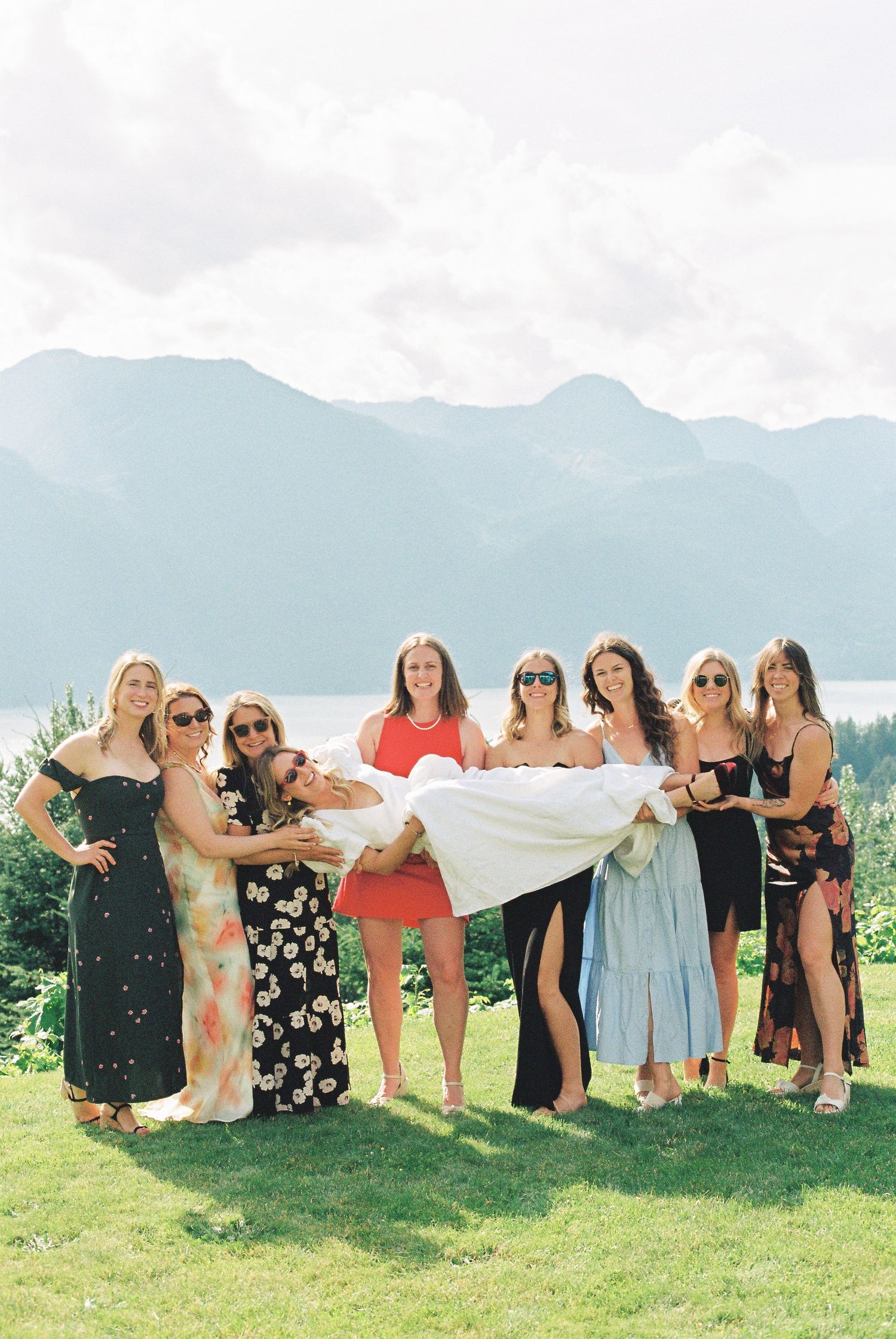 Bridesmaids lifting and carrying smiling bride outdoors with stunning mountain and lake backdrop.