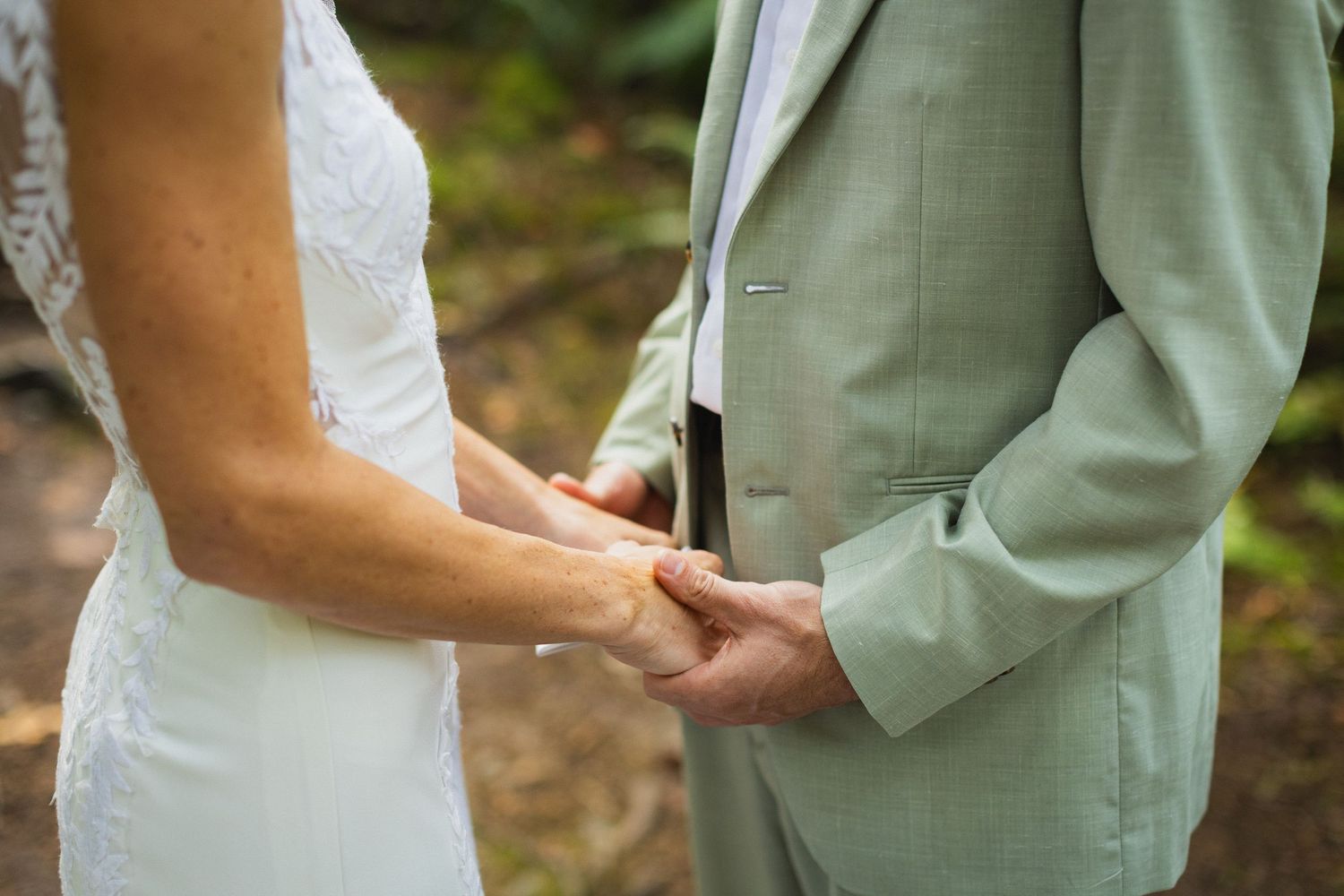 Close-up of bride and groom holding hands during wedding ceremony in sage green suit.