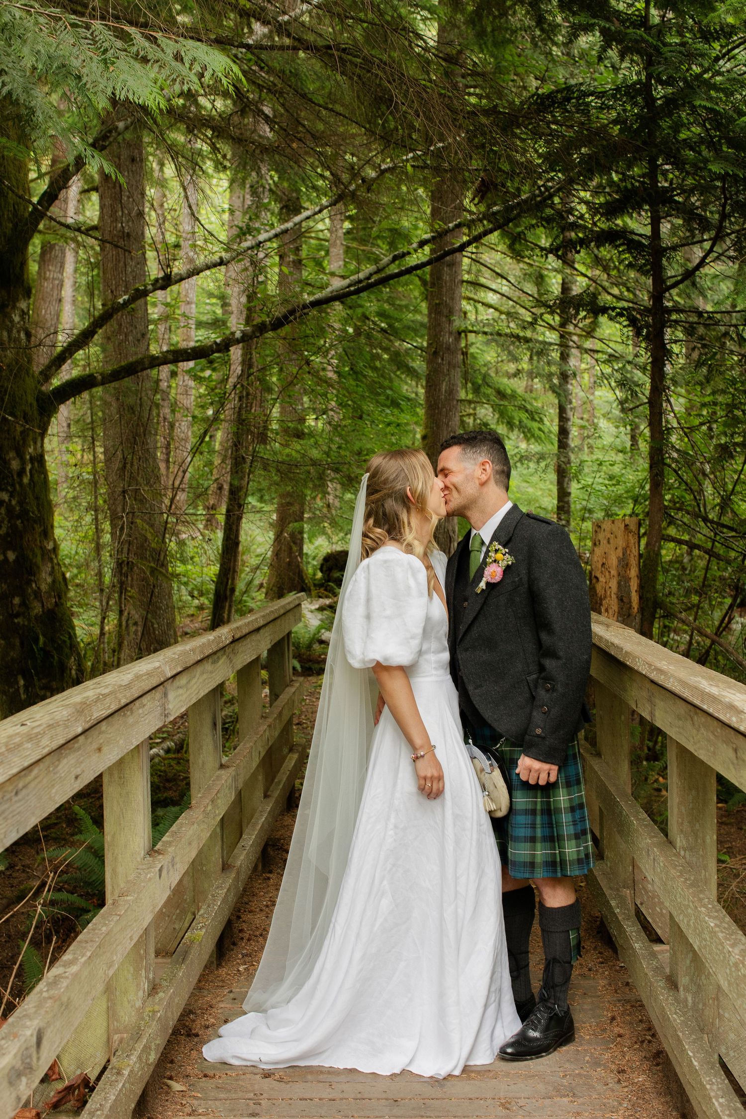 Bride and groom sharing a kiss on a wooden forest bridge, groom wearing a traditional Scottish tartan kilt.