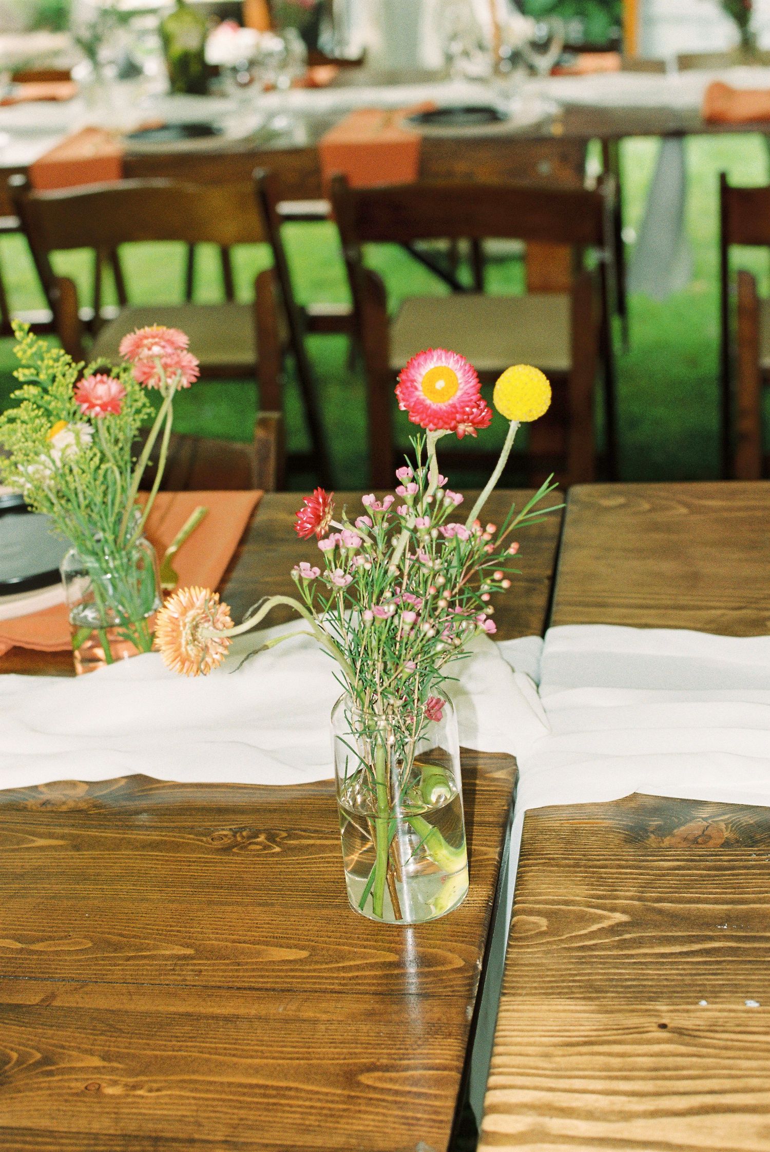 Rustic wooden wedding reception table decorated with colorful wildflower centerpieces in glass vases.