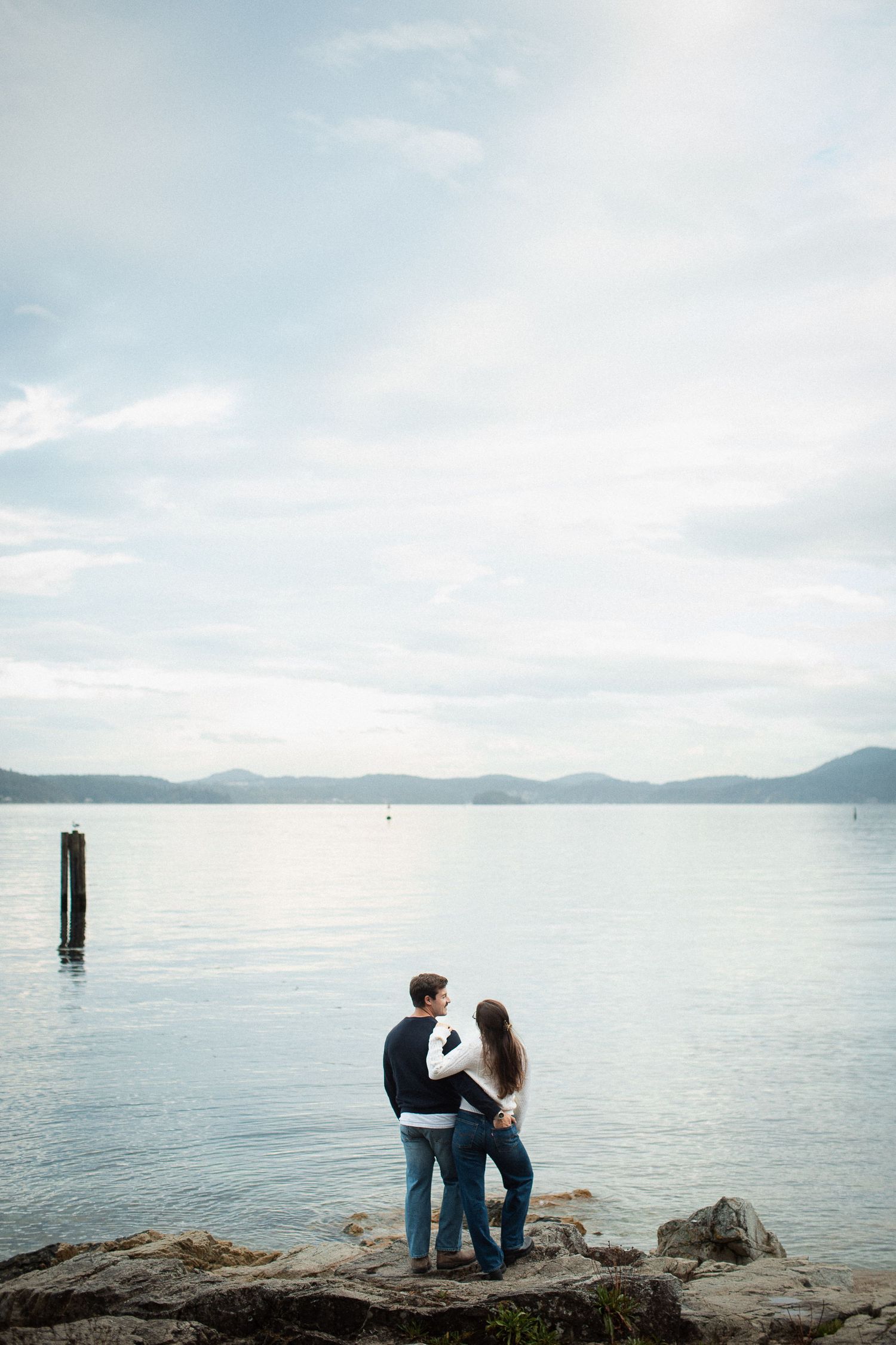 Couple standing together on rocky shore gazing at a calm lake with mountain views.