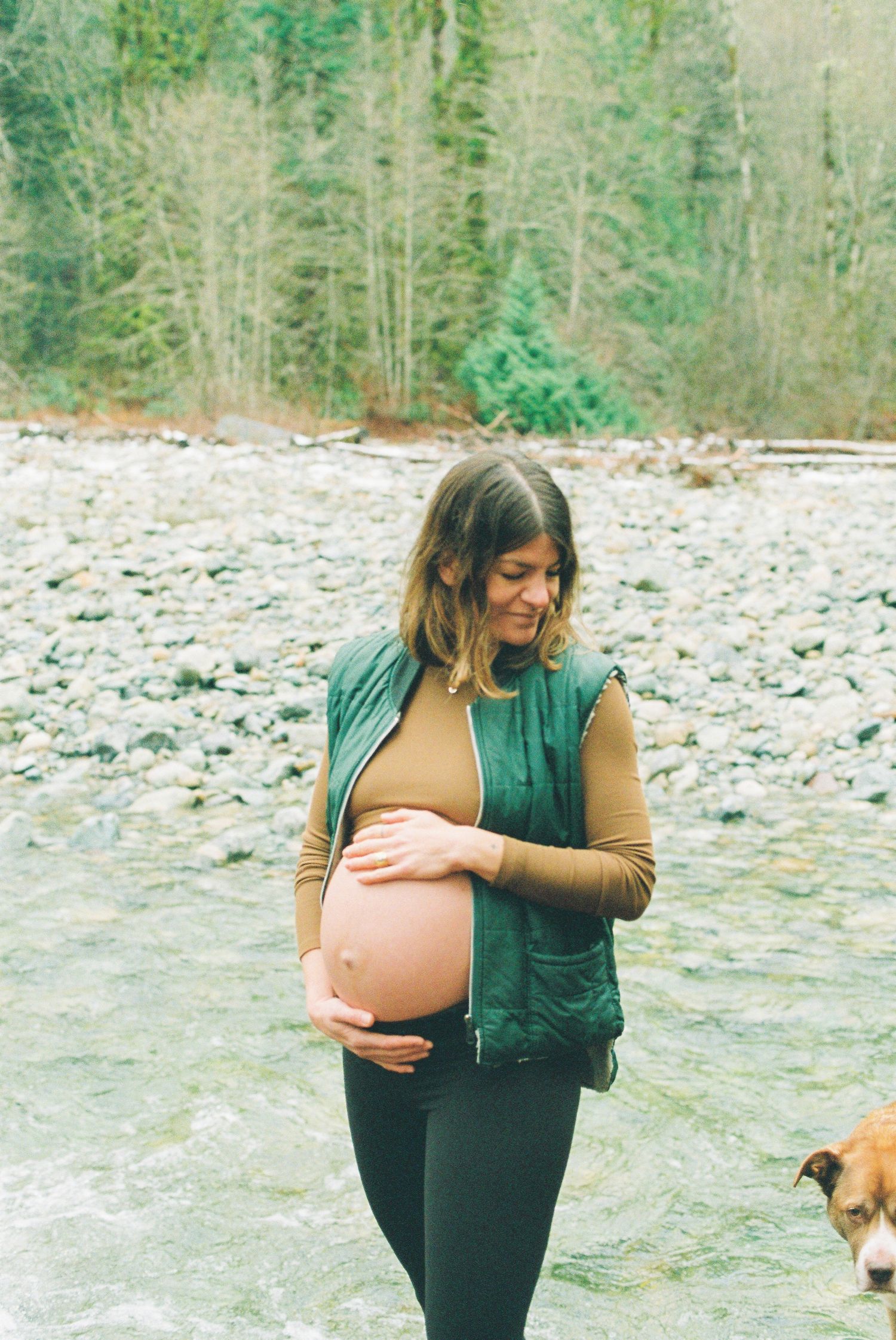 Pregnant woman in green vest tenderly holding baby bump by rocky mountain river.