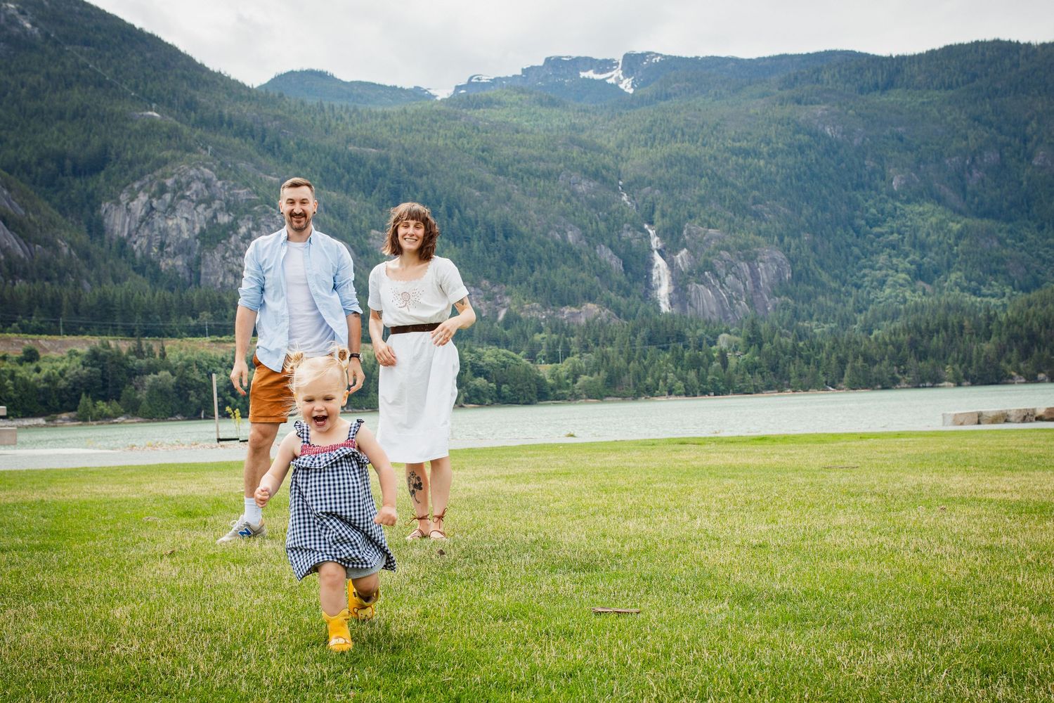Family of four with toddler running ahead on green grass with scenic mountain waterfall backdrop.