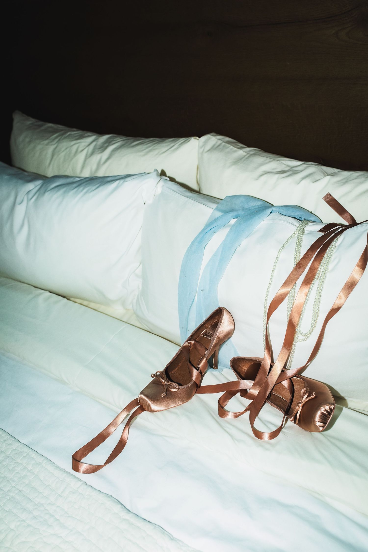 Satin bridal heels with ribbon ties resting on white hotel bed pillows.