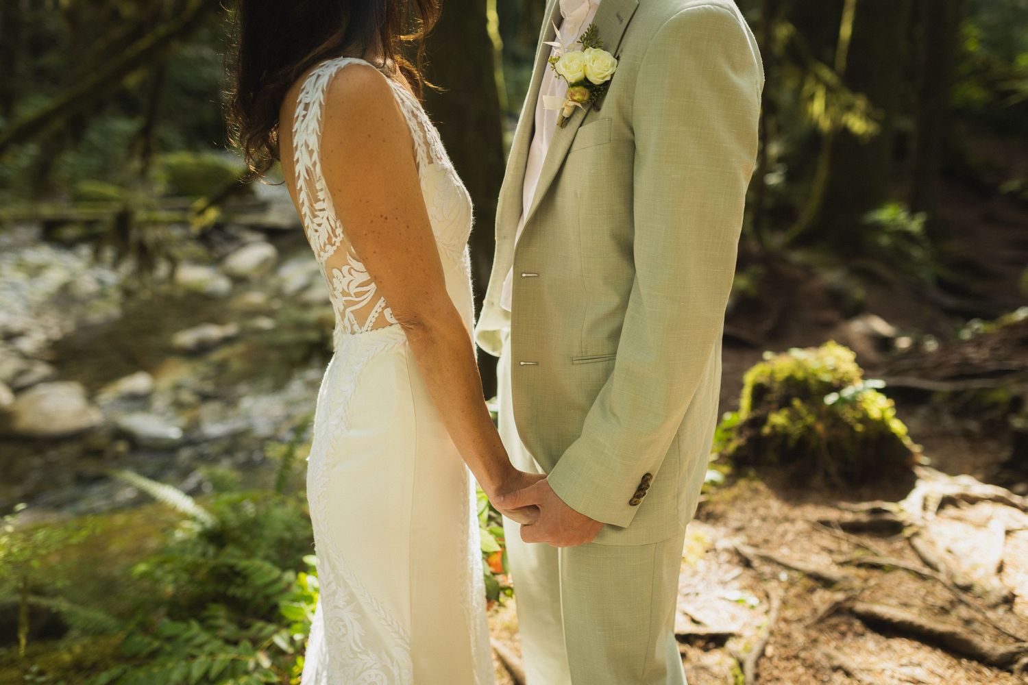 Close-up of bride and groom holding hands in front of a mossy forest stream backdrop.