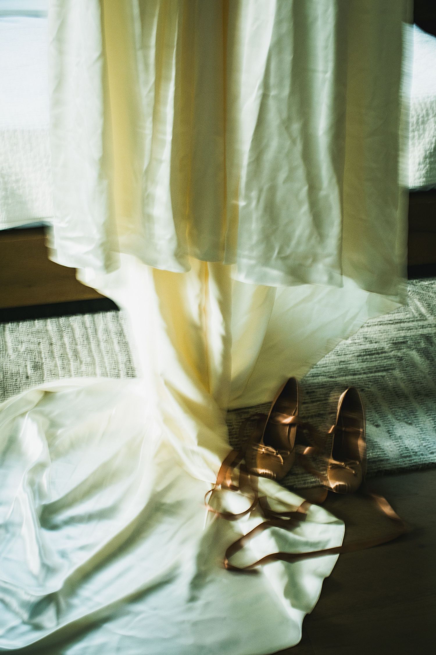 Gold bridal heels with ribbon ties resting on white wedding dress near sunlit sheer curtains.