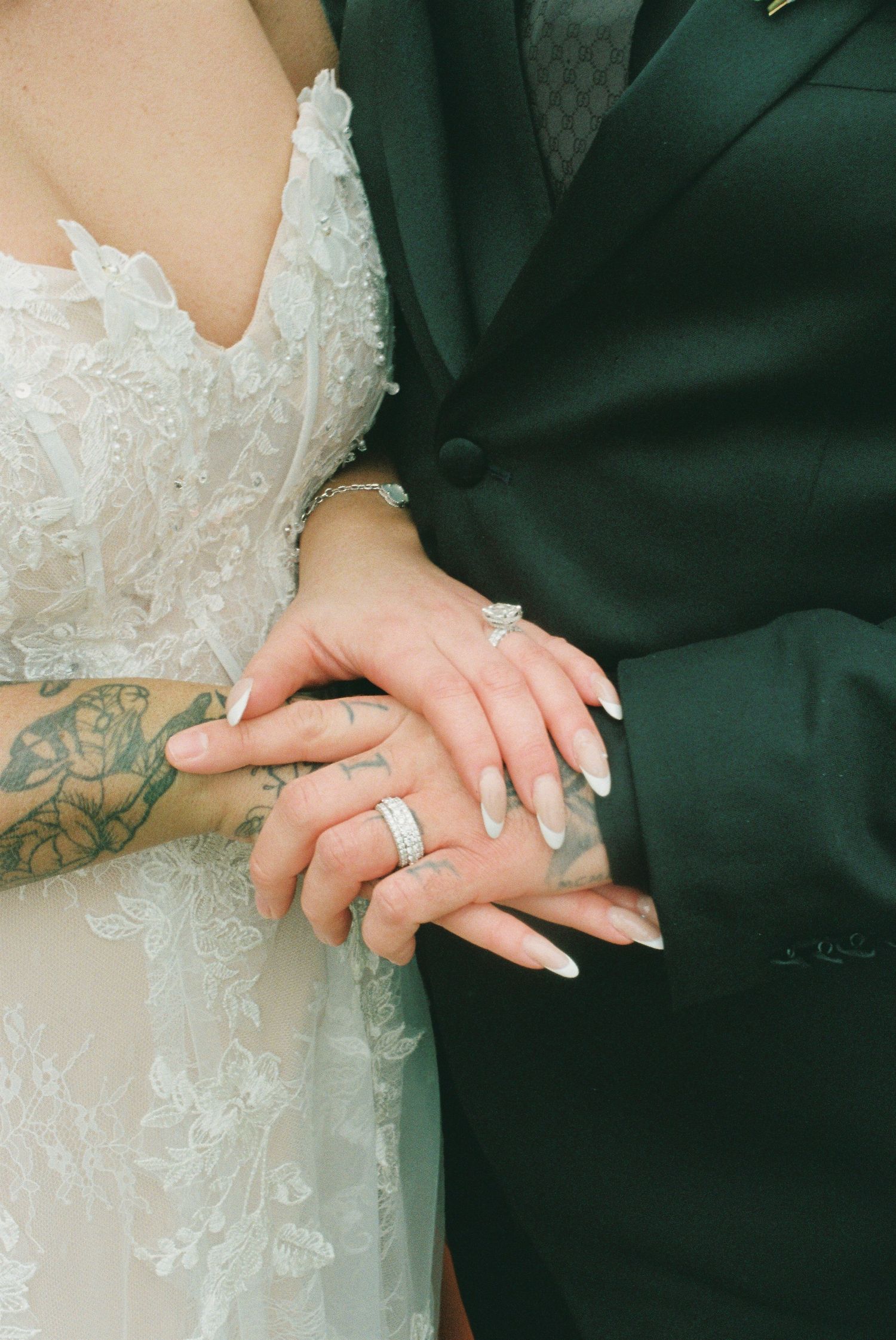 Close-up of bride and groom hands showing wedding rings, tattoo, and white nails.