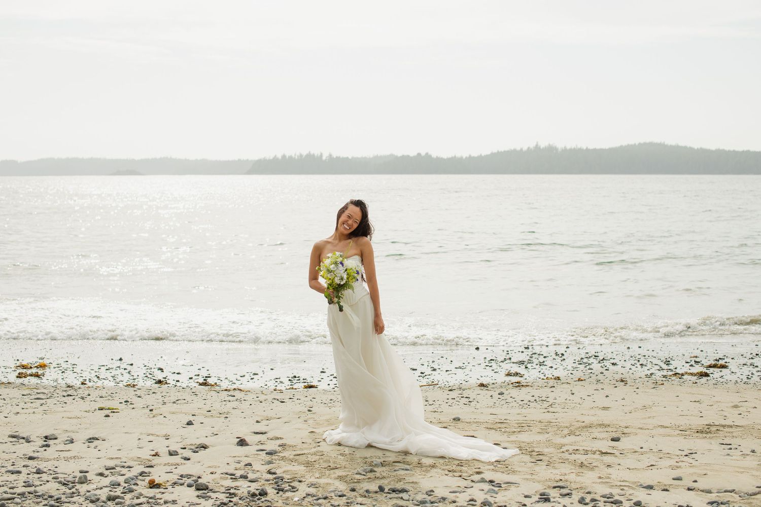 Bride in white gown holds bouquet while standing alone on a shimmering sandy beach by the ocean.
