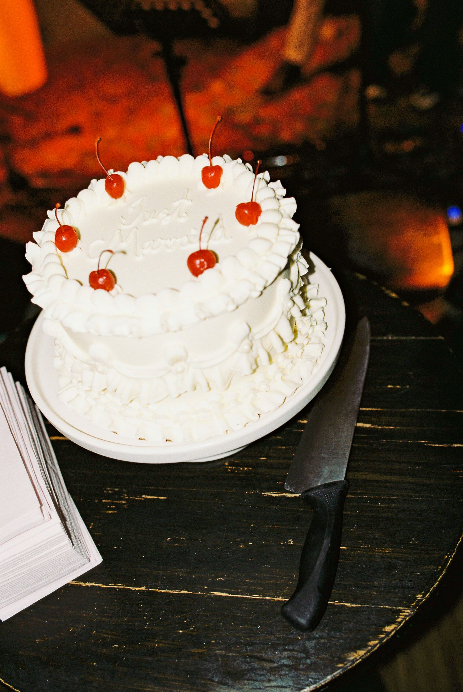 Two-tier white wedding cake decorated with red cherries on a dark wooden table.