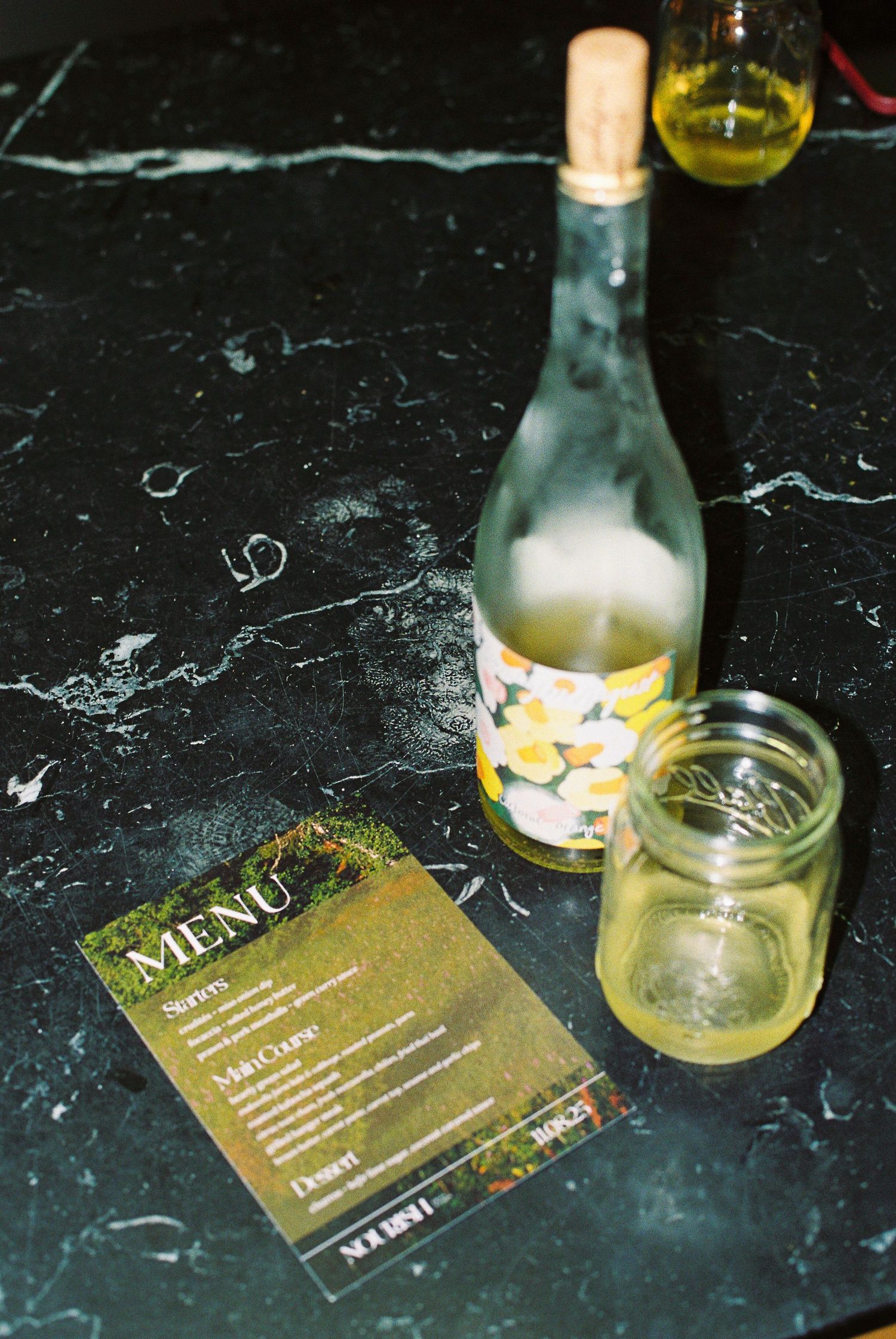 Wine bottle with floral label, mason jar glass, and dinner menu card arranged on dark marble surface.