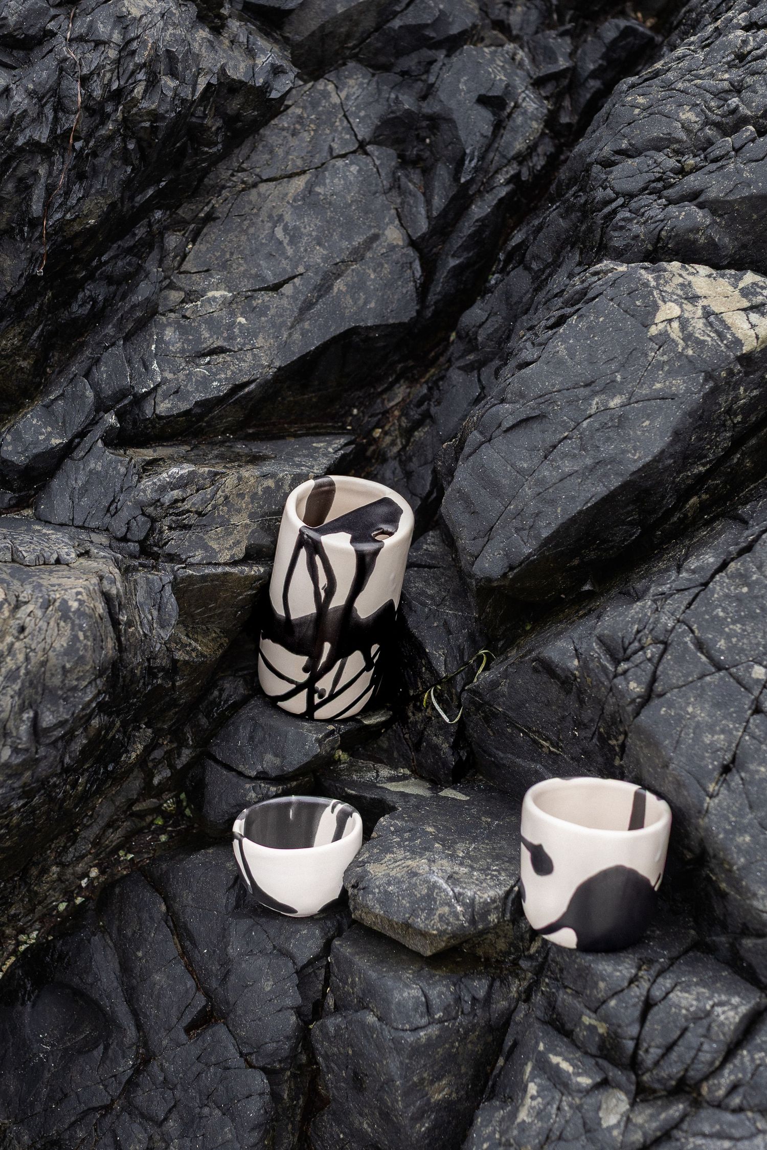 Black and white ceramic cups and vase arranged on dark rocky coastal surface.