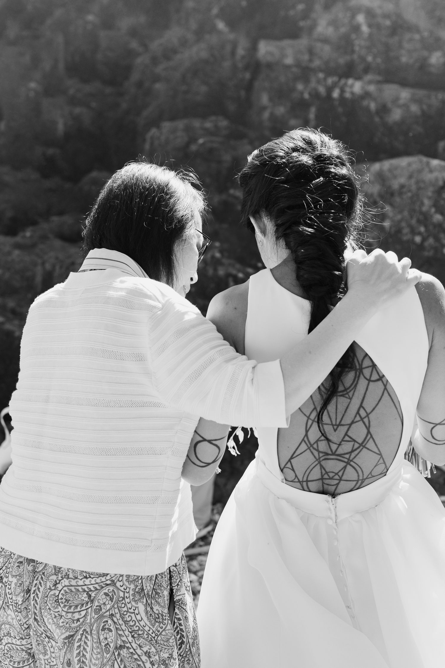 Black and white photo of older woman adjusting tattooed bride's wedding dress from behind outdoors.
