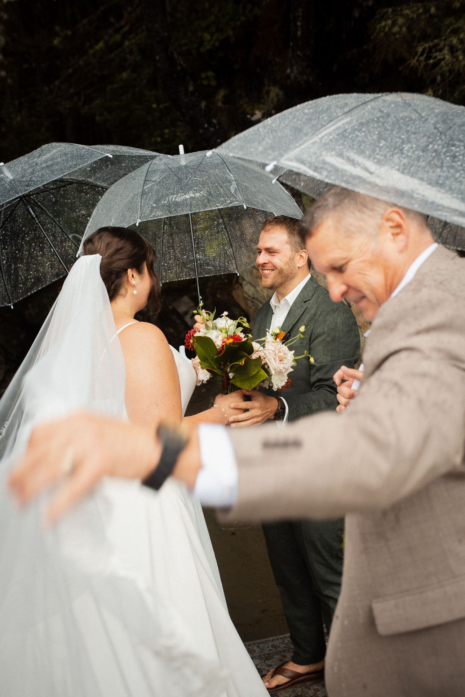 Bride and groom exchange vows under clear umbrellas in a rainy outdoor wedding ceremony.