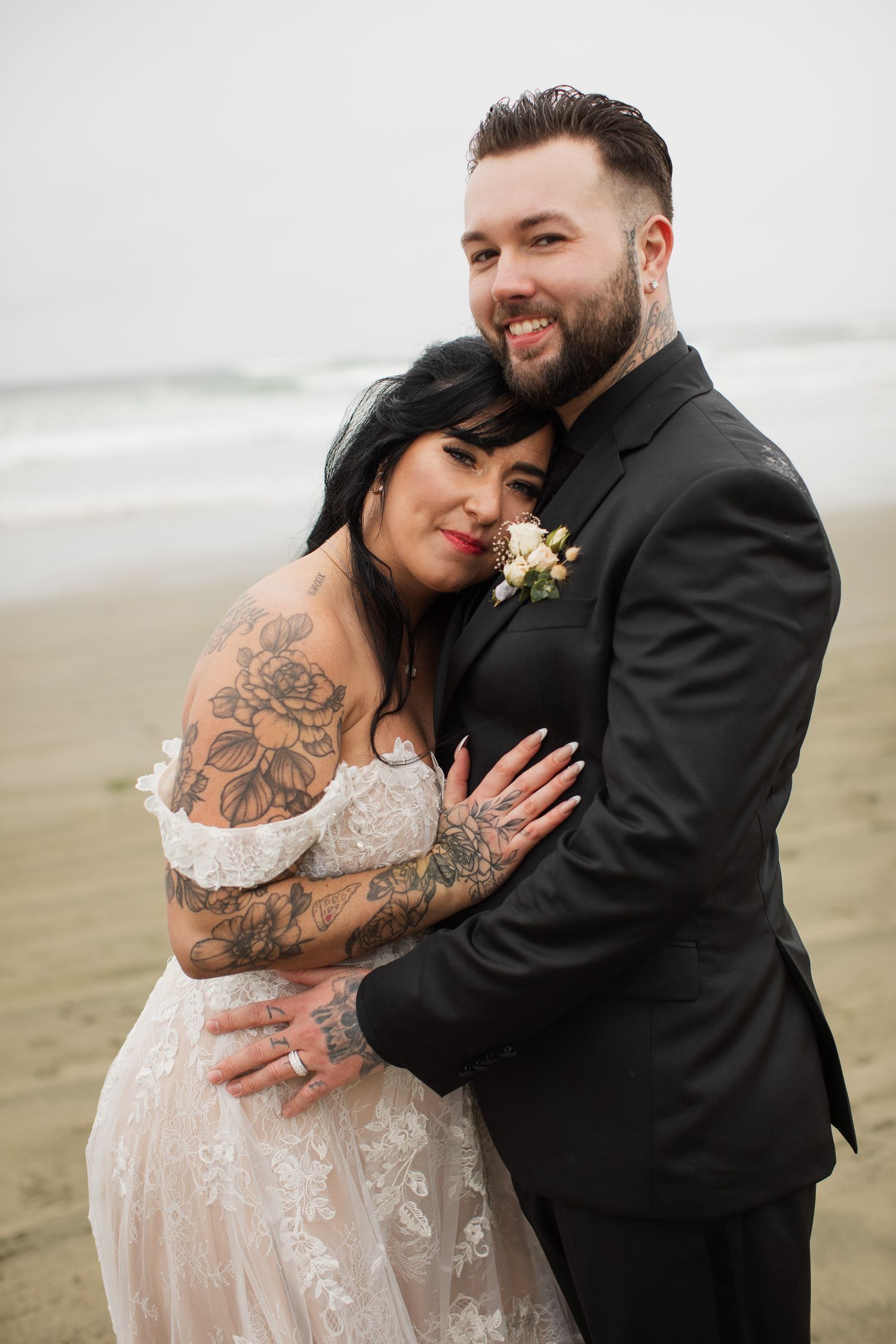 Tattooed bride in lace gown leans into smiling groom in black suit on overcast beach.
