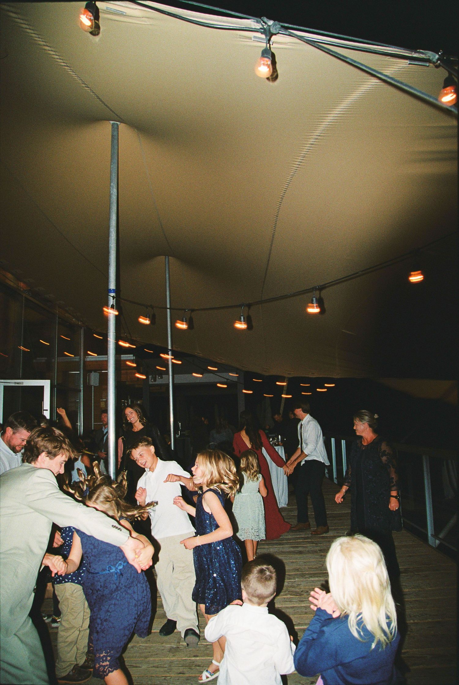 Wedding guests dance energetically under a large tent with string lights at lively mountain reception.