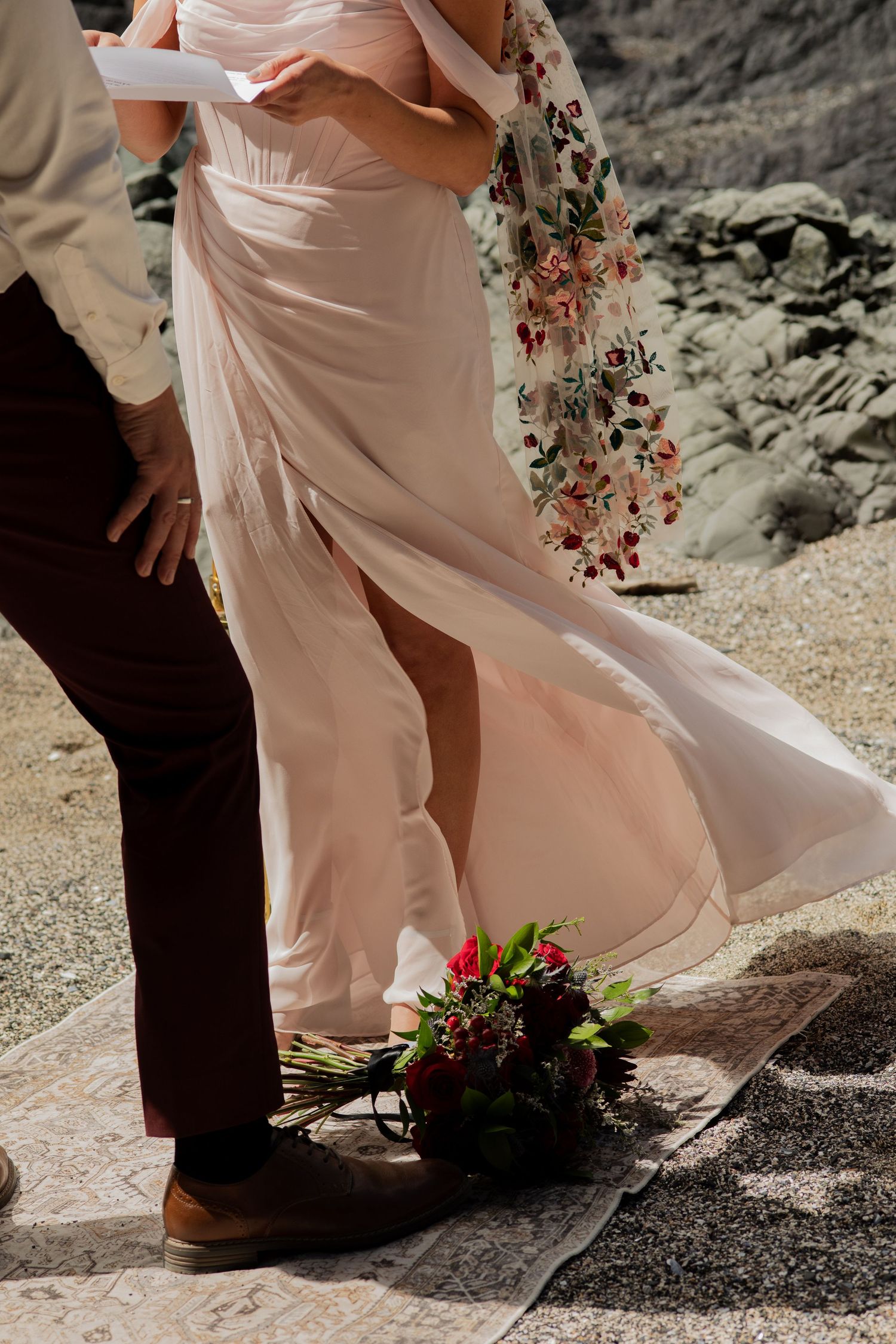 Close-up of bride in blush pink dress with floral veil and red bouquet at beach wedding.
